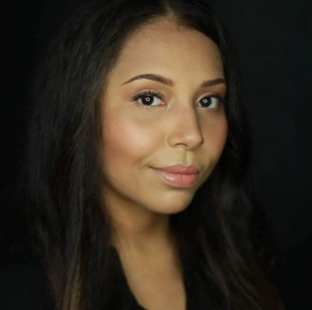 A close-up of a person with long, dark hair smiling and looking at the camera against a black background.