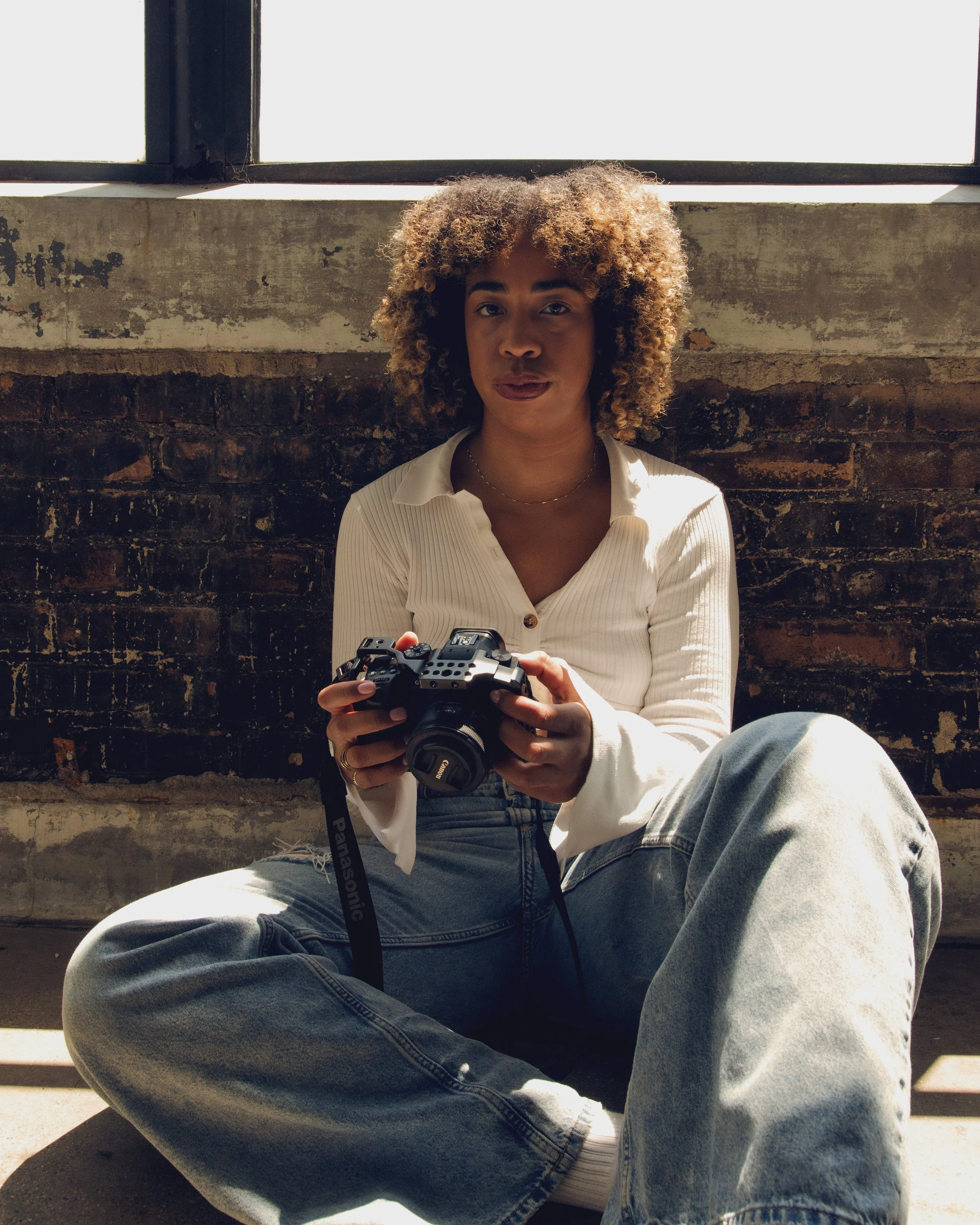 Person with curly hair sitting on the ground in front of a brick wall, holding a camera, near a large window.