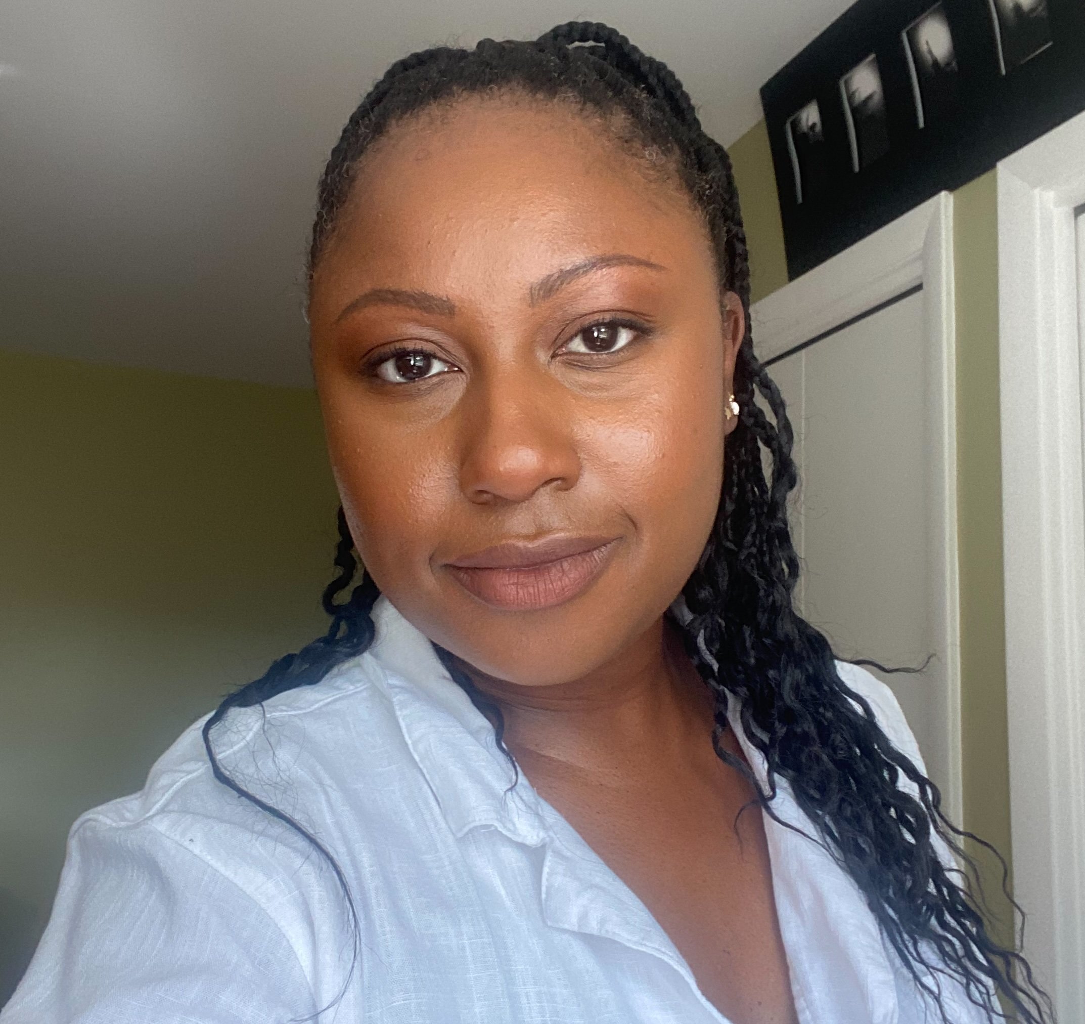 Close-up of a person's face showing their eyes, forehead, and smile, with braided hair and earrings, in a well-lit room with a white door in the background.