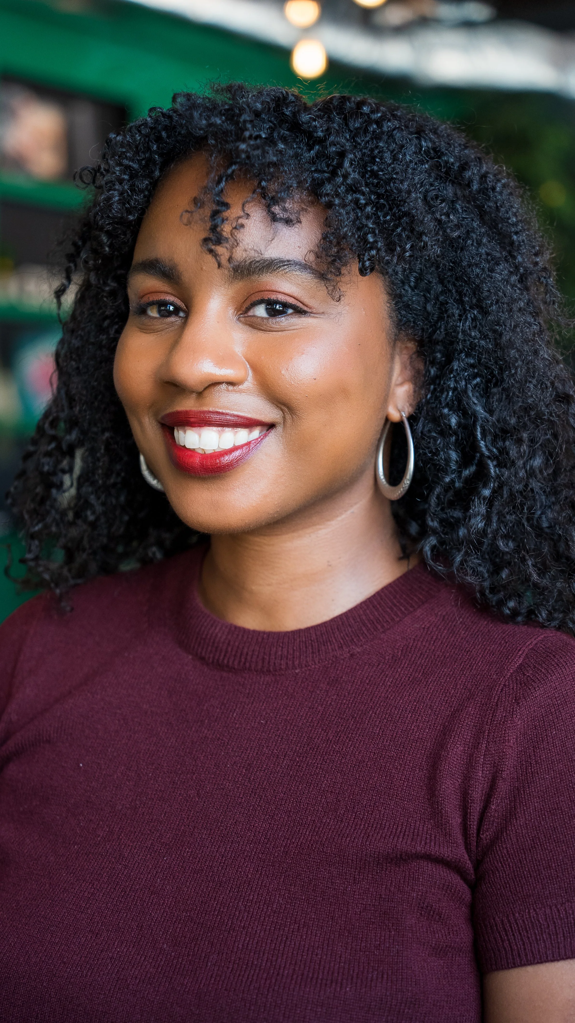 Close-up of a woman with curly black hair wearing a maroon top and hoop earrings, smiling in an indoor setting.