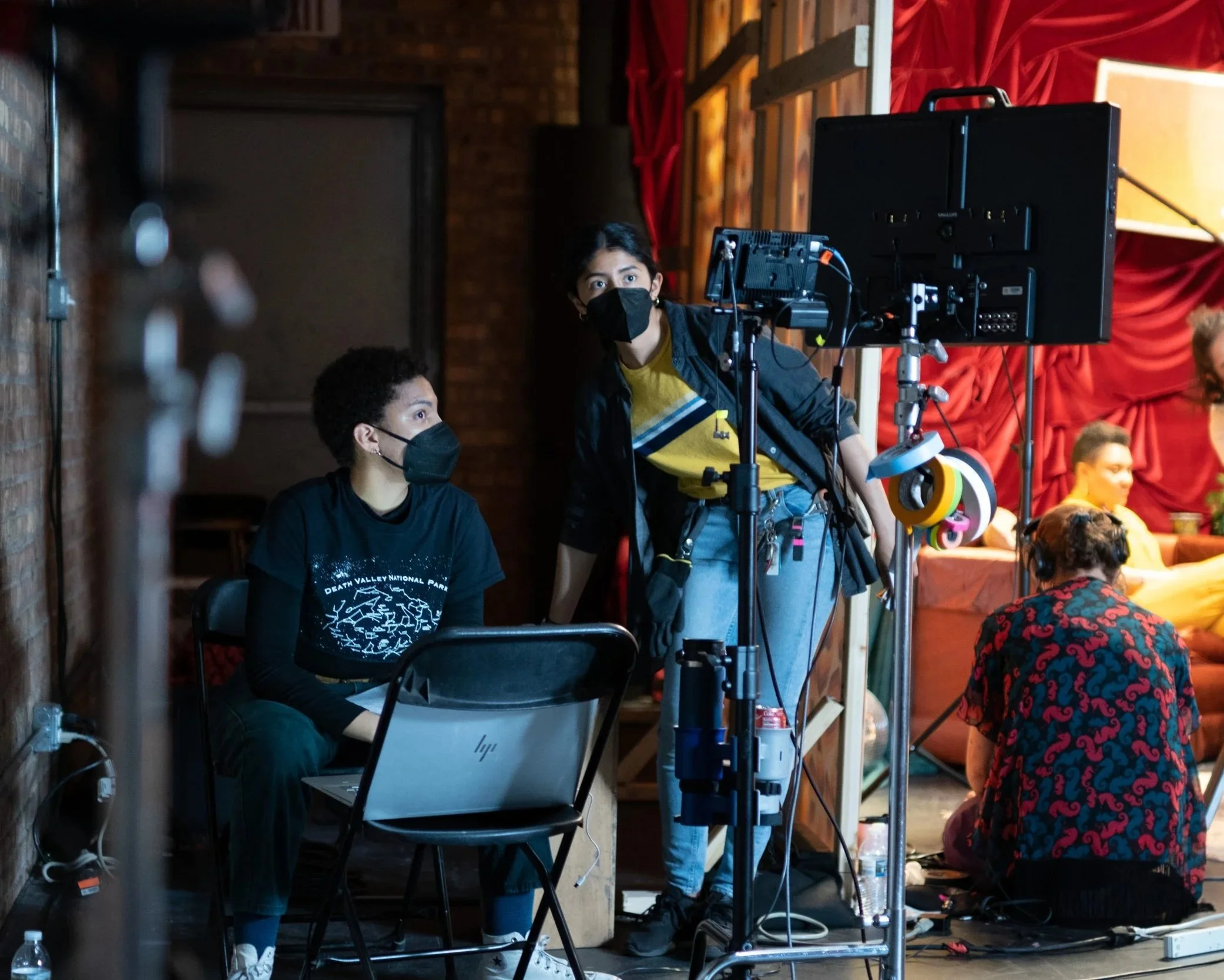 A group of people working with filming equipment indoors, including a person operating a camera and others sitting around with masks on.