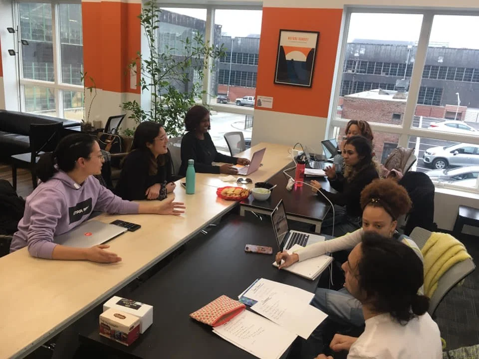 A group of people gathered around a conference table in a modern office, engaged in a discussion or meeting, with laptops, notebooks, and a pizza on the table, and large windows in the background.