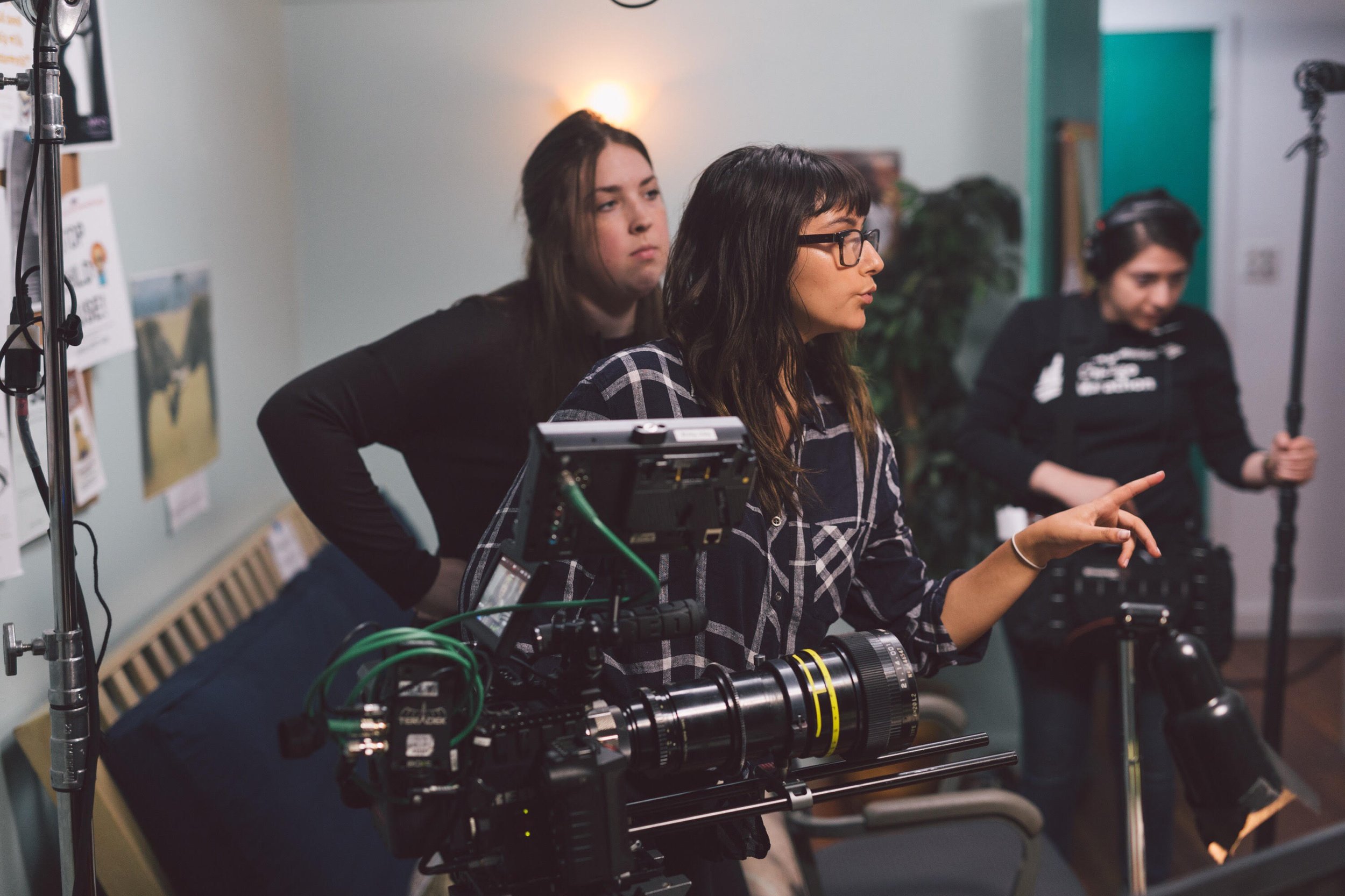 Three people working on a film or video production set. One person with glasses points at equipment, while another with long hair looks on, and a third person in the background wears headphones and operates a camera.