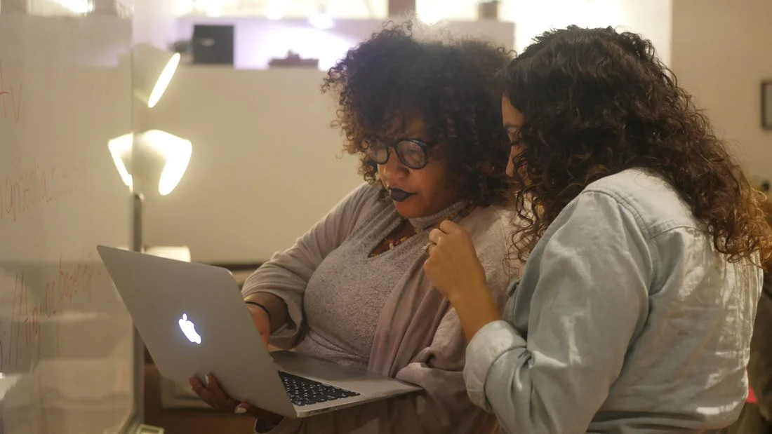 Two women with curly hair and glasses looking at a laptop together in an office setting.
