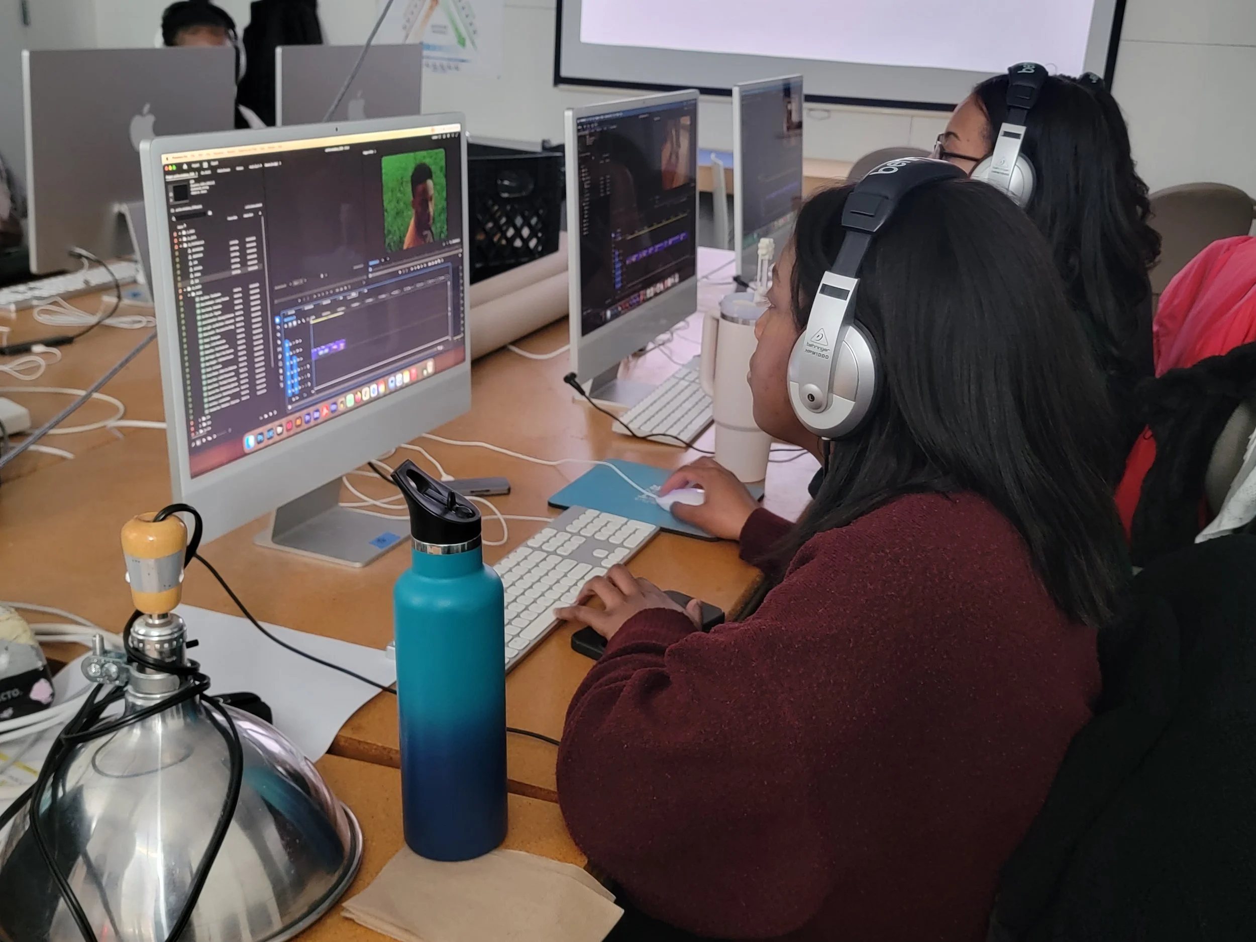 Two people working on video editing at computers in a classroom setting, with video editing software open on the screens.