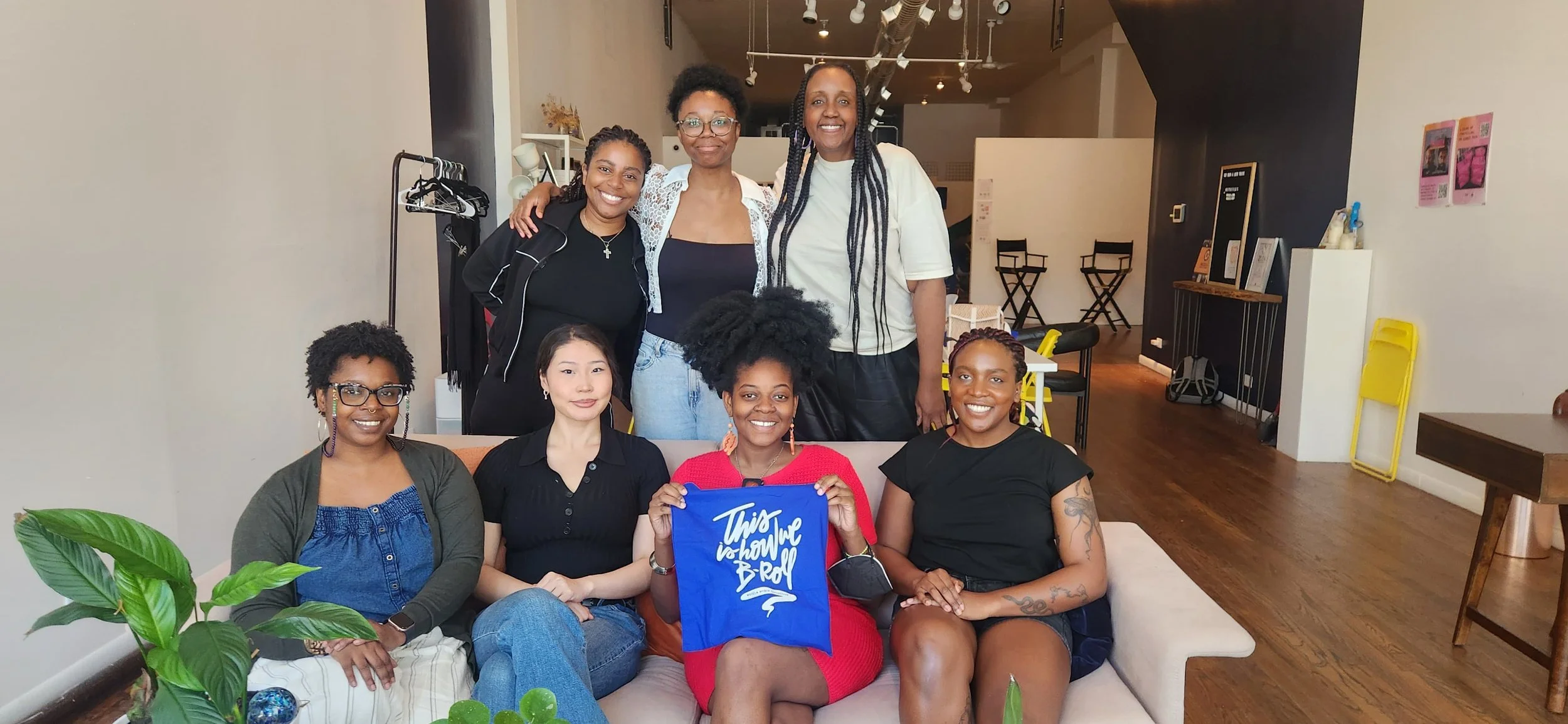 A group of nine smiling and posing together in a cozy, modern indoor space. Some are seated on a sofa, and others are standing behind it. One is holding a blue bag with white text that says 'This is how we B-Roll.' The background includes artwork, chairs, and a clothing rack.
