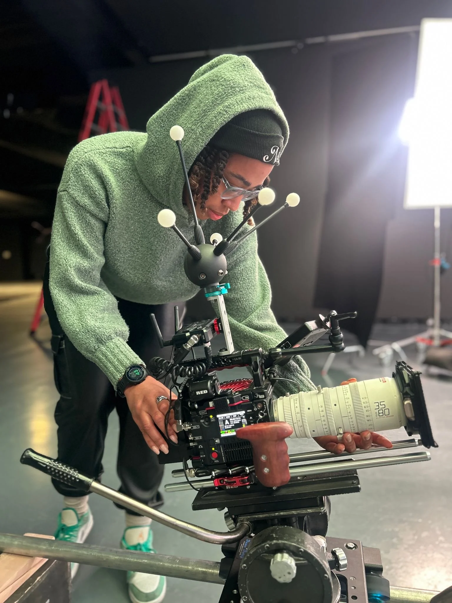 Person operating professional video camera mounted on a rig, wearing a green hoodie, black beanie, and glasses, in a studio setting.