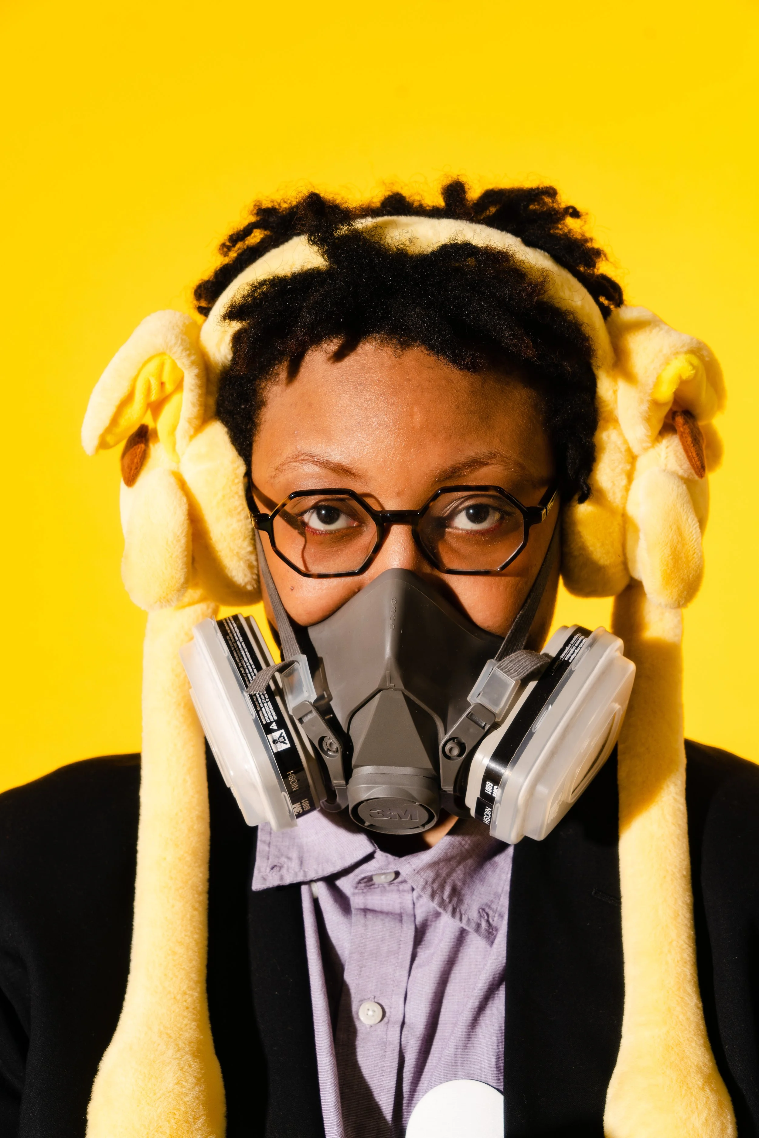 A person with glasses wearing a yellow headband with plush animal ears, a mask, and a respirator, against a yellow background.