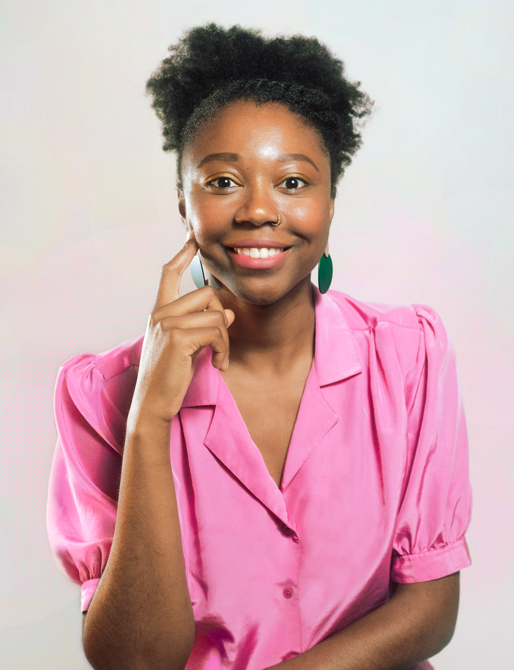 A person with dark skin and natural hair styled in a puff, wearing a pink satin blouse with puffed sleeves and green earrings, smiling and touching their cheek with their finger.