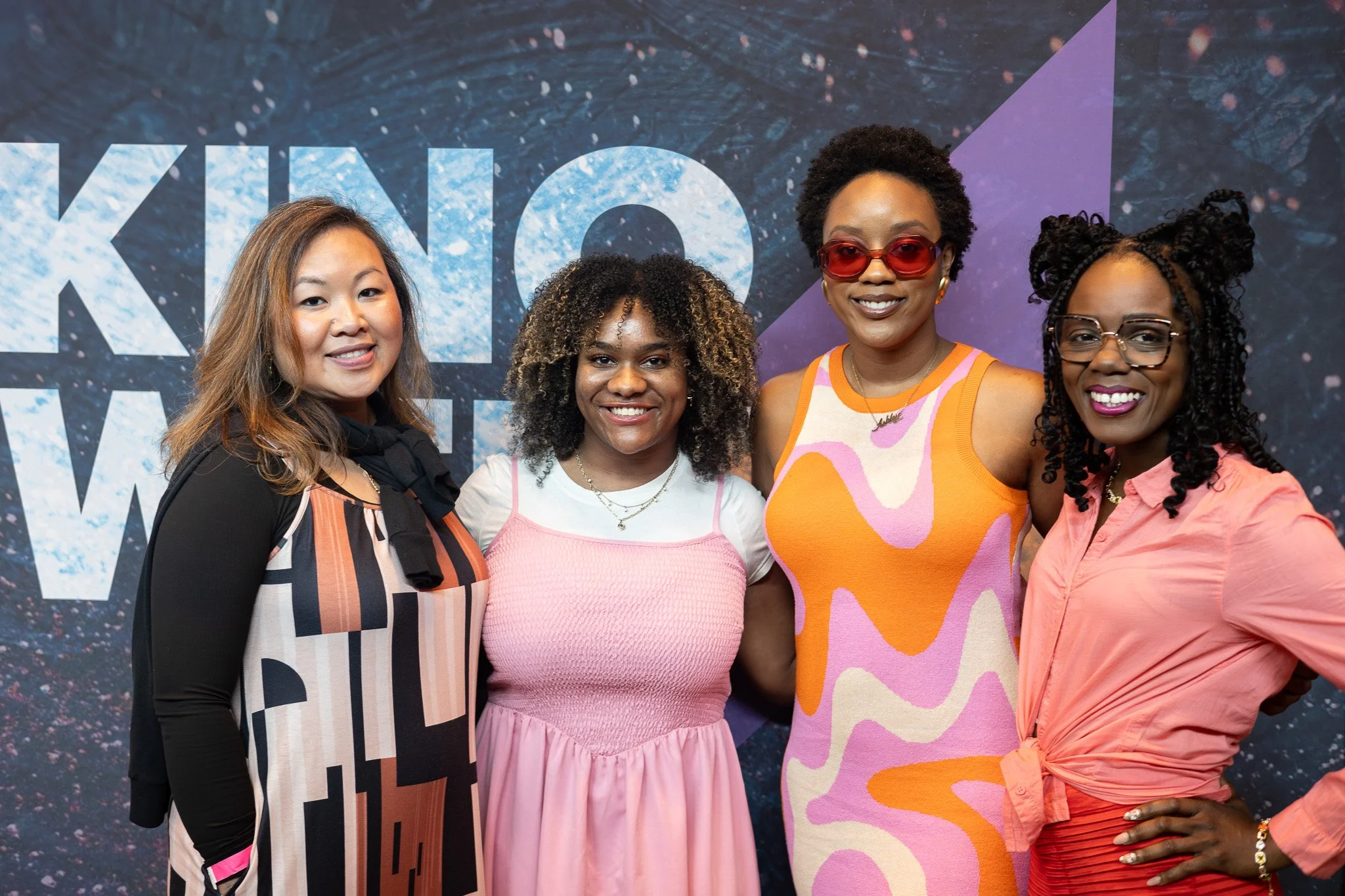 Four people standing together and smiling in front of a dark cosmic-themed background. They are dressed in colorful, trendy outfits with accessories, and appear to be at a public or promotional event.