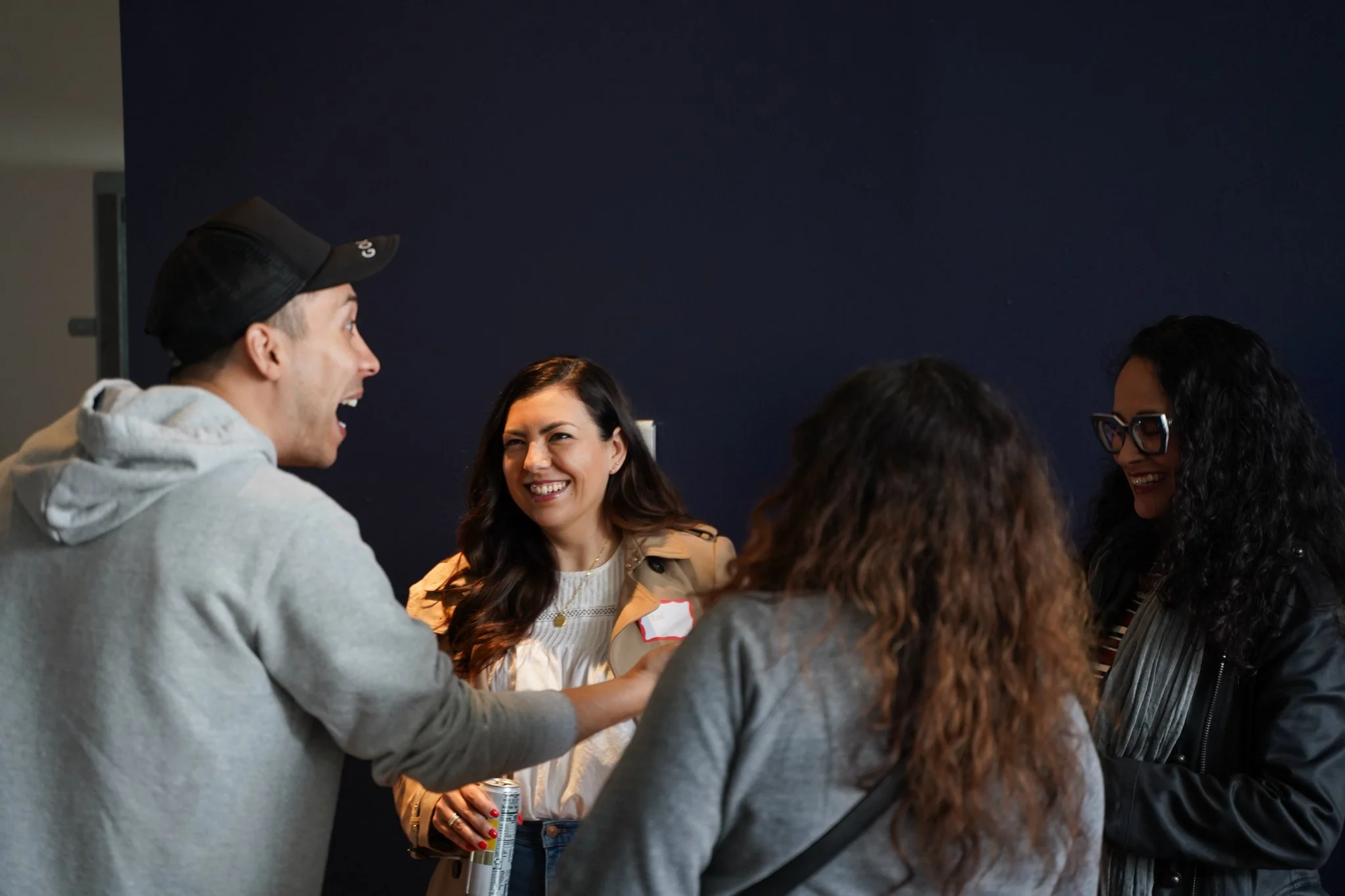 A group of people engaged in conversation, smiling and laughing in an indoor setting with a dark blue wall.