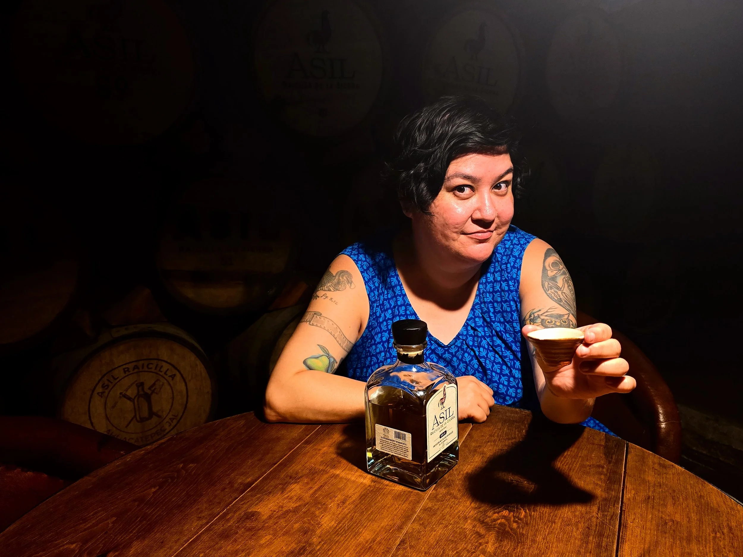 A woman with dark, short hair and tattoos on her arms is sitting at a wooden table, holding a small wooden bowl. She is wearing a blue sleeveless top with a leaf pattern and has a bottle of liquor with a label on the table. The background features barrels stacked against a dark wall with faint circular logos.