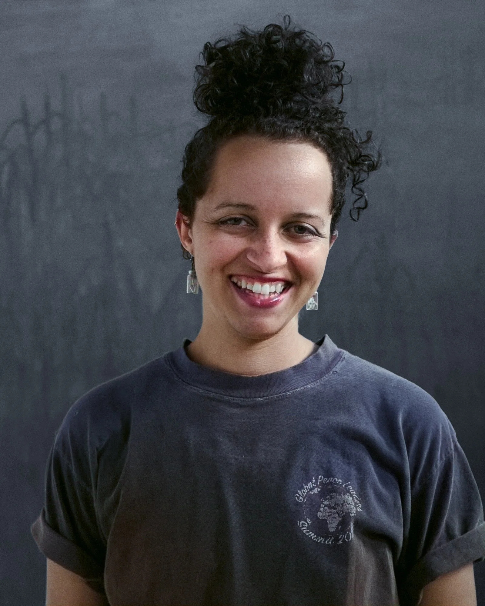 Close-up of a person with natural curly hair styled in a high bun, smiling against a dark background.