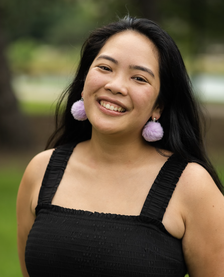 A smiling person with long black hair wearing a black dress and purple fluffy pom-pom earrings outdoors with blurred trees in the background.