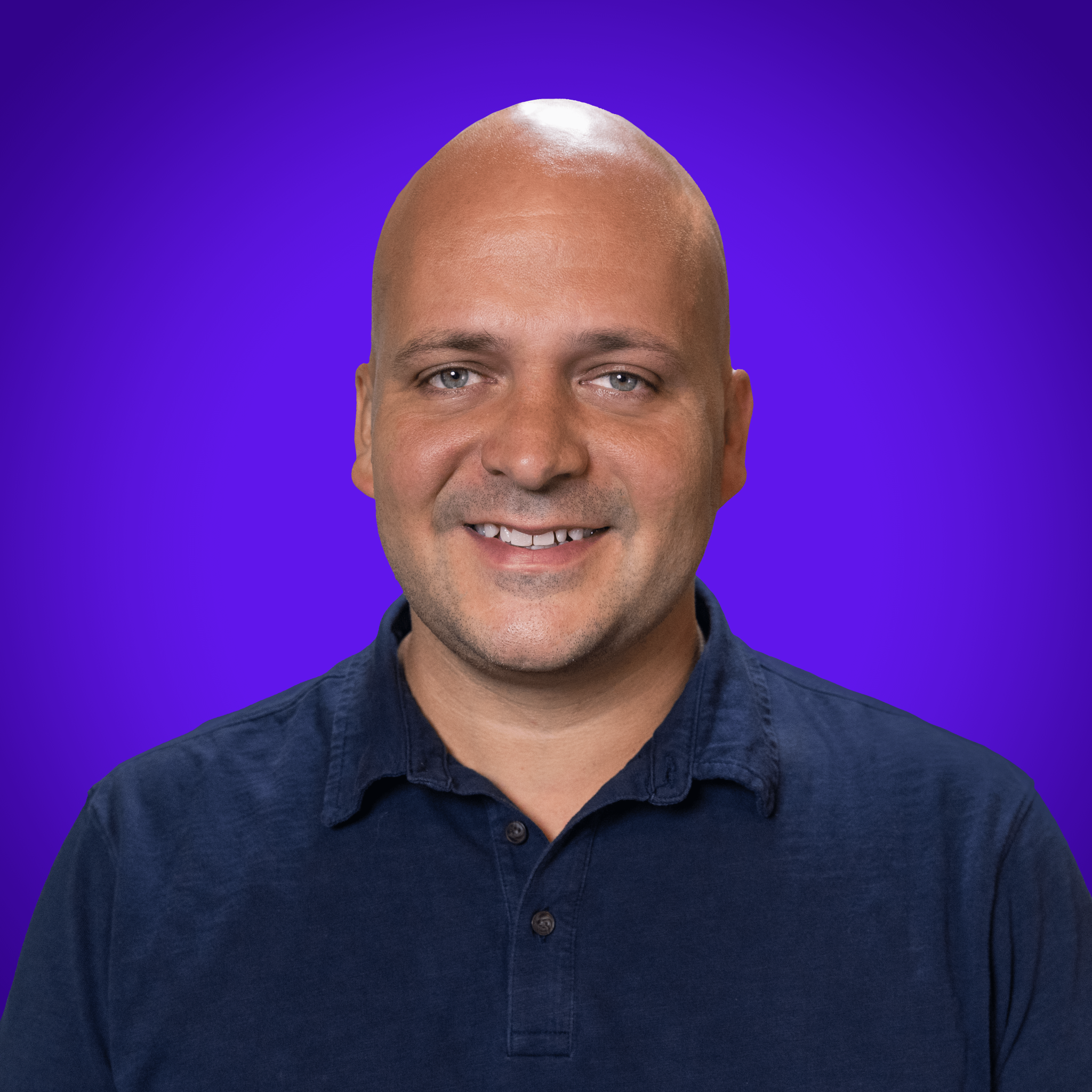 Close-up of a smiling man with light blue eyes and a shaved head, wearing a dark blue shirt, against a vibrant purple background.