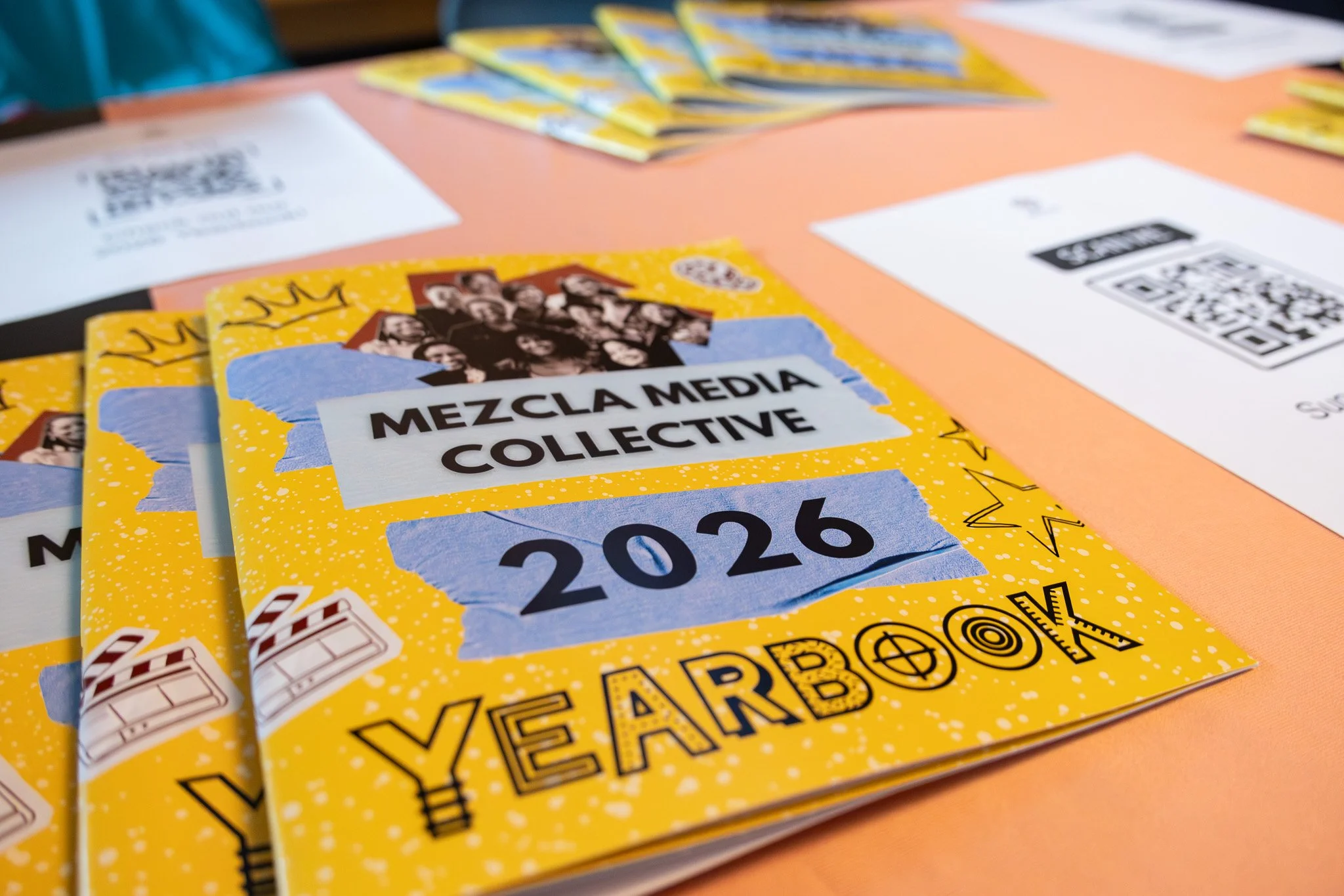 Fliers with a yellow background, featuring a group photo, text saying "Mezcla Media Collective," and the year 2026, placed on a table with QR codes and documents.