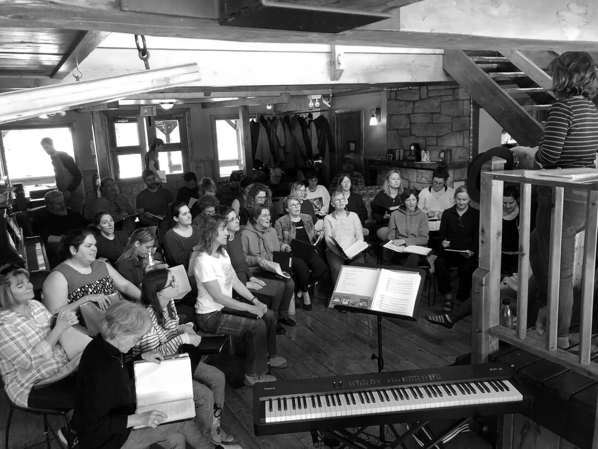Un groupe de personnes assises dans une pièce, probablement en train de chanter ou d'écouter une répétition, avec un piano électrique au premier plan et un chef d'orchestre ou de chorus à droite, en train de diriger.