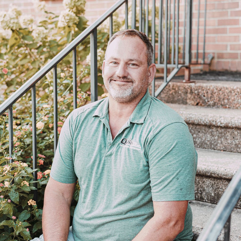 A middle-aged man with short hair, beard, and mustache sitting on steps outside, wearing a light green polo shirt with a logo, smiling at the camera. Background includes brick wall and plants.