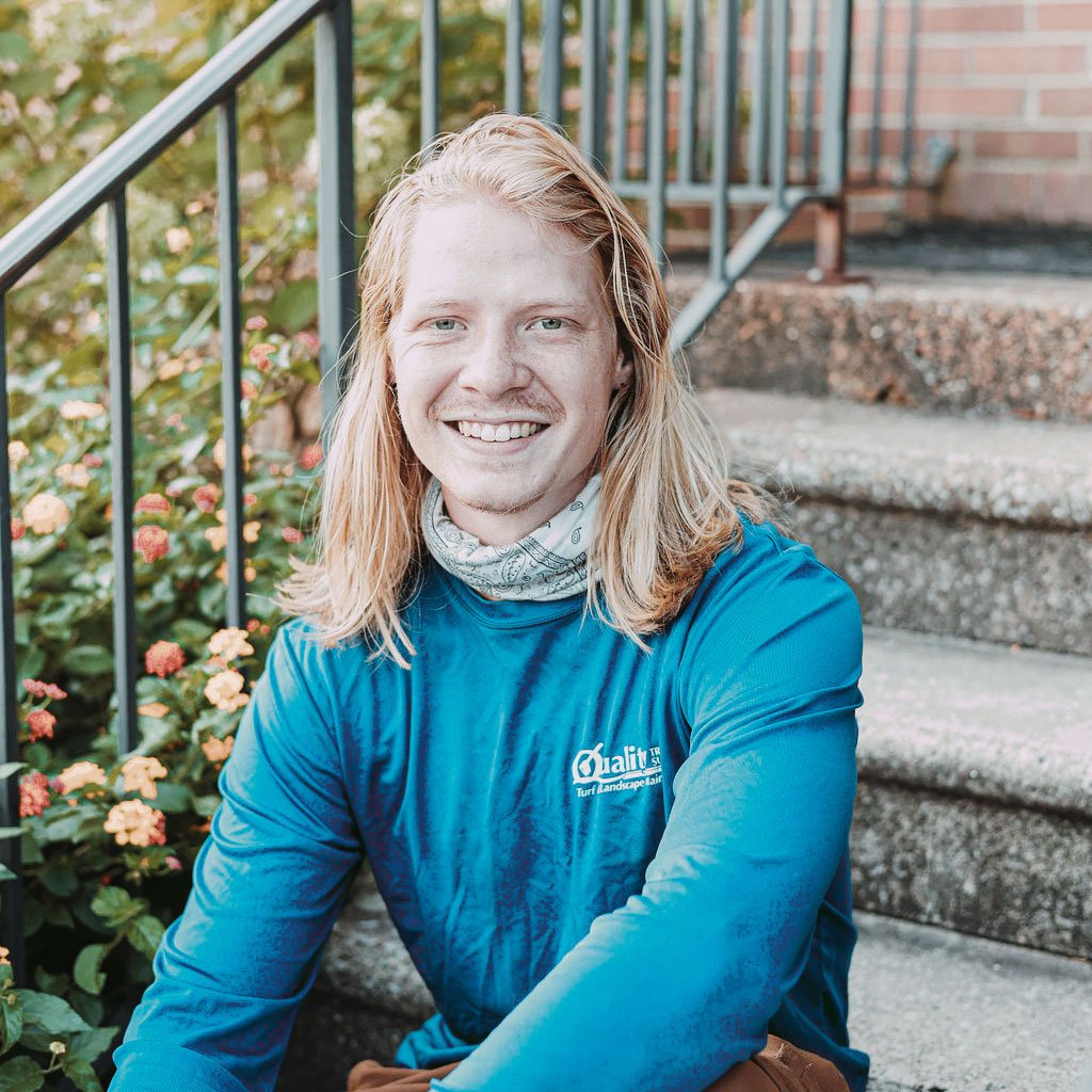 A young man with long blond hair, smiling, sitting on outdoor stairs, wearing a blue long-sleeve shirt and a white bandana around his neck, with a brick building and green plants in the background.