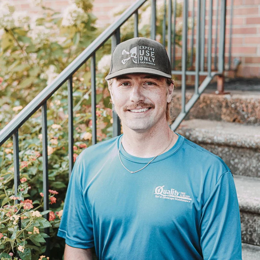 A man with a mustache and blonde hair wearing a blue T-shirt and a baseball cap, standing outdoors near a brick building and a staircase with plants in the background.
