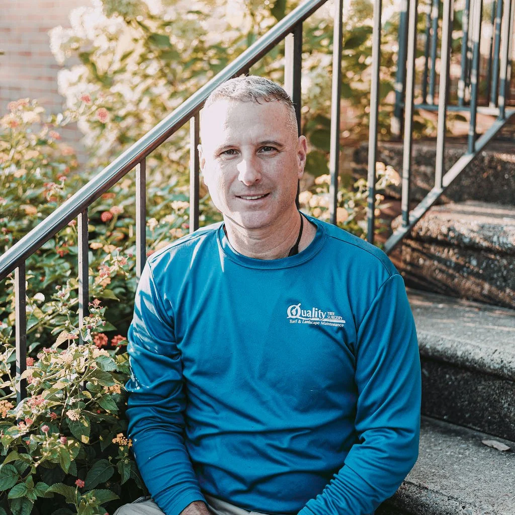A man sitting on outdoor steps near a garden with pink flowers, wearing a blue long-sleeve shirt with a logo that reads 'Quality Tree Surgery Turf & Landscape Maintenance'.