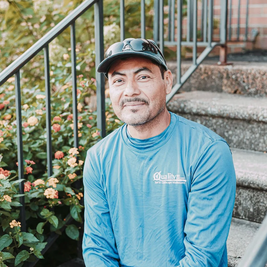 A man wearing a blue long-sleeve shirt and sunglasses sitting outdoors on concrete stairs, with a black metal railing and green bushes with orange flowers in the background.