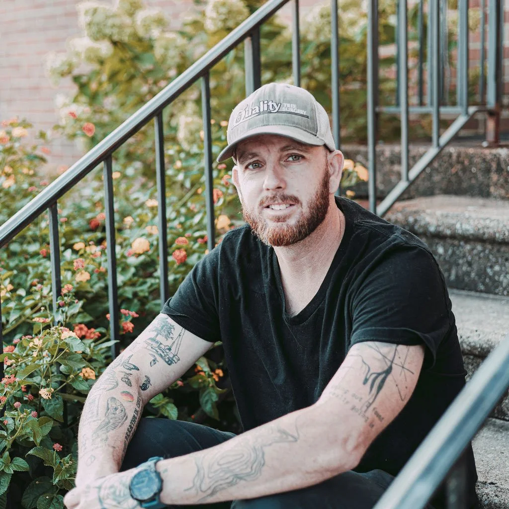 A man with a beard and tattoos wearing a gray cap and black t-shirt sitting on outdoor steps next to a garden with green plants and orange flowers.