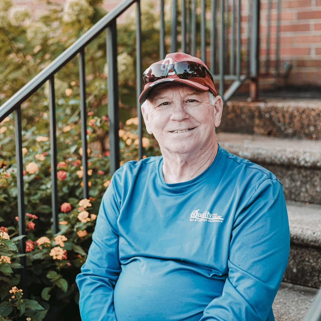An elderly man sitting on outdoor stairs next to a flower bed, wearing a red baseball cap, sunglasses on the cap, a blue long-sleeve shirt, and smiling.
