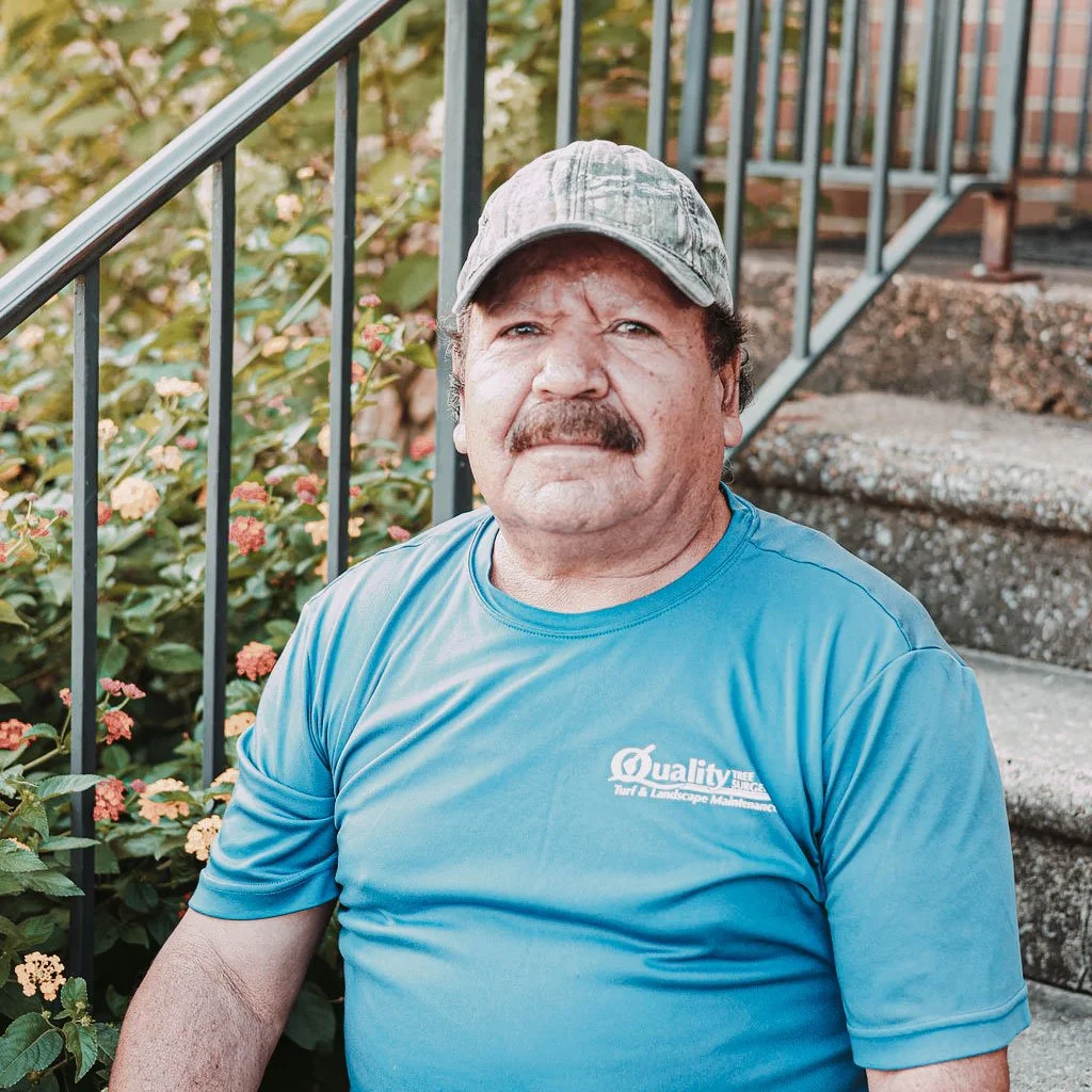 A man with a mustache wearing a blue T-shirt and a gray cap sitting outdoors on a set of concrete stairs next to a black metal railing and pink flowering bushes.