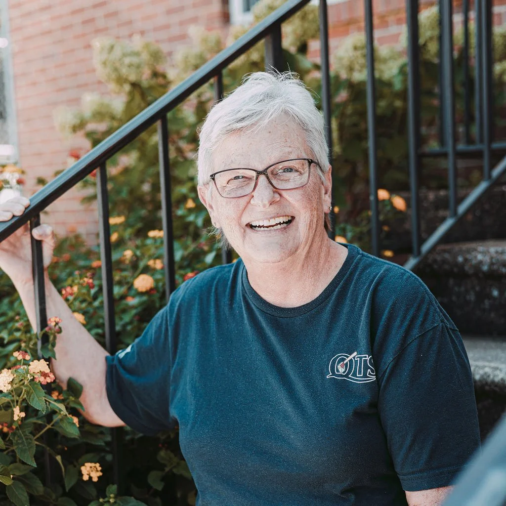 An elderly woman with gray hair and glasses smiling and holding a black metal railing outside a house with brick walls and plants.