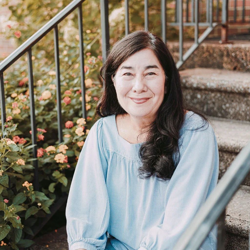 A woman with long dark hair smiling, sitting on outdoor stairs next to a garden with pink and orange flowers.