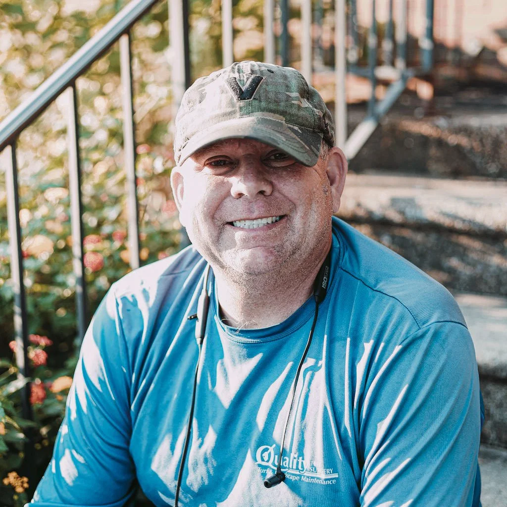 A smiling man wearing a camouflage cap and a blue shirt sitting on outdoor stairs with greenery in the background.