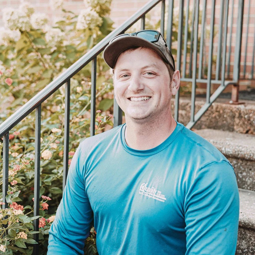 A smiling man wearing a blue long-sleeve shirt, a baseball cap, and sunglasses on his cap, sitting on outdoor steps near a railing with a garden of green plants and flowers behind him.