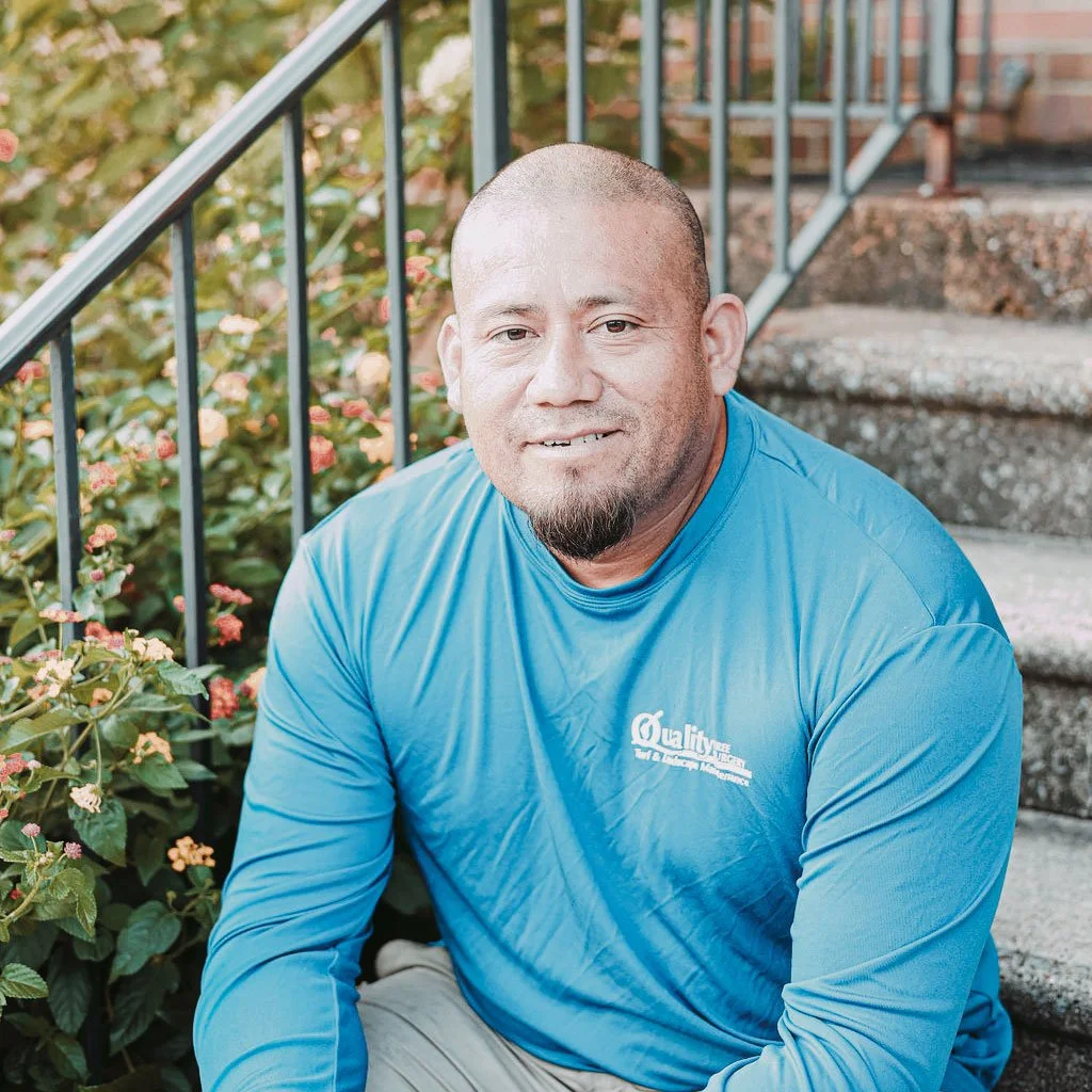 A man with a shaved head and goatee sitting on outdoor stairs next to flowering bushes, wearing a blue athletic shirt.