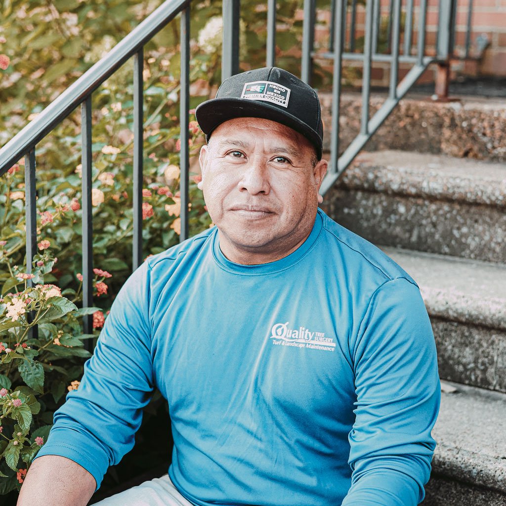 A man sitting outdoors on concrete steps, wearing a blue shirt and a black cap, with plants and a metal railing in the background.