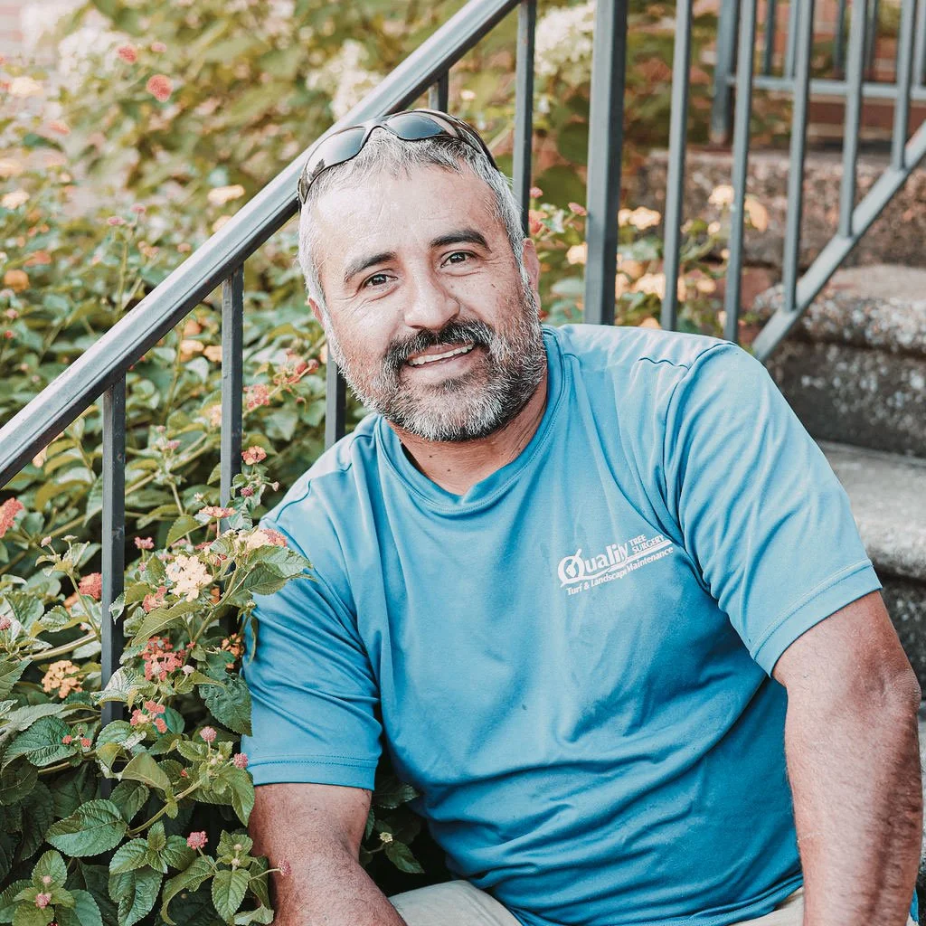 A middle-aged man with gray hair and beard, wearing a blue T-shirt and sunglasses on his head, sitting on outdoor stairs next to green bushes and flowers, smiling at the camera.