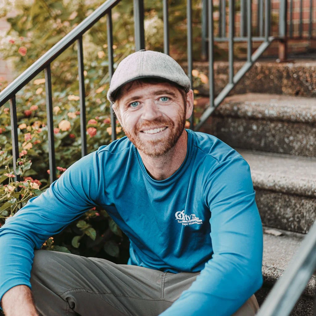 A smiling man with a beard and blue eyes, wearing a gray flat cap and a blue long-sleeve shirt, sitting on outdoor concrete stairs with a railing and greenery in the background.