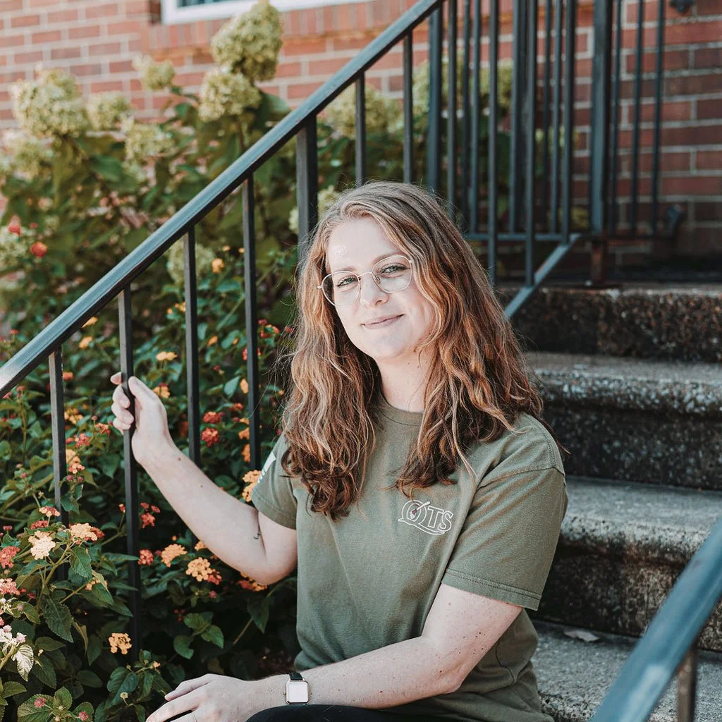 A young woman with curly brown hair and glasses sitting on outdoor stairs next to flowering bushes, smiling at the camera.