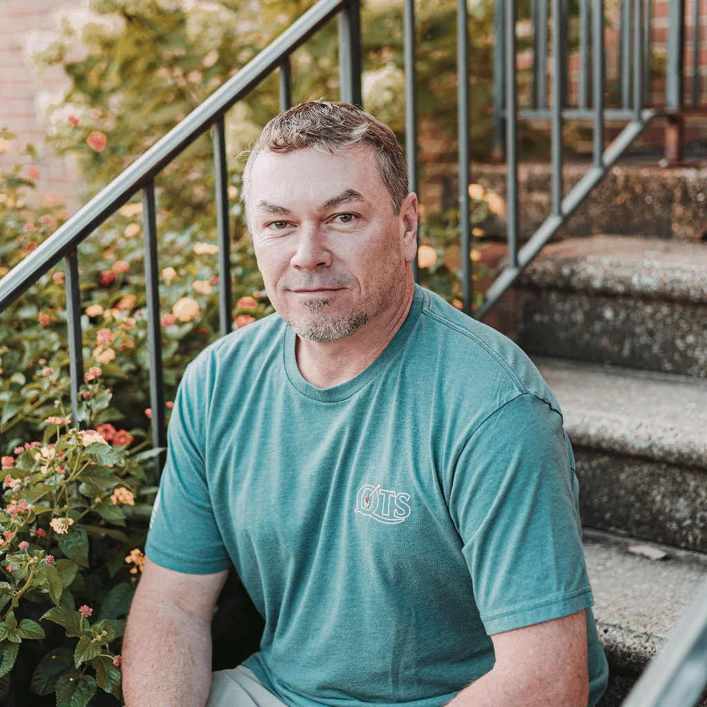 A man sitting on outdoor concrete stairs next to flowering plants, wearing a teal T-shirt and looking at the camera.