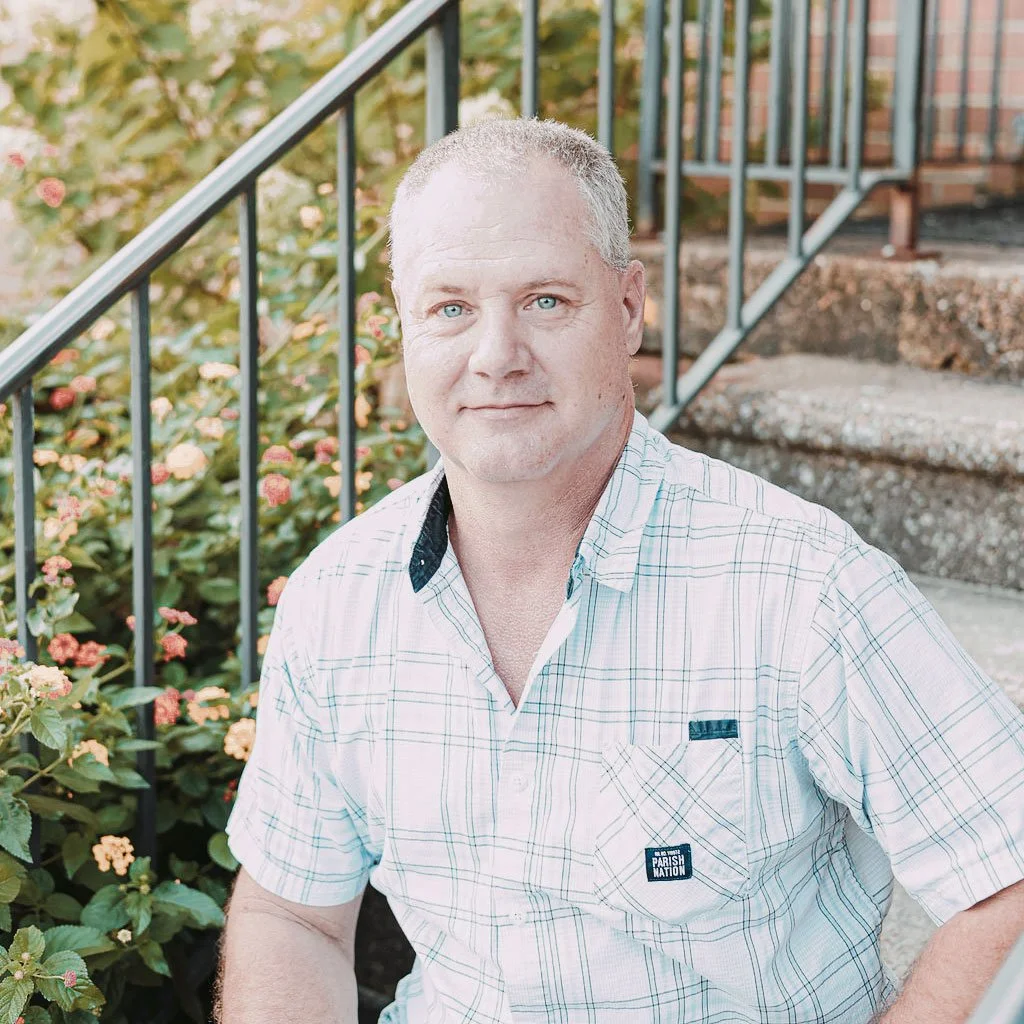 A middle-aged man with short gray hair and blue eyes, wearing a plaid shirt, sitting outdoors near a staircase with green plants and flowers in the background.