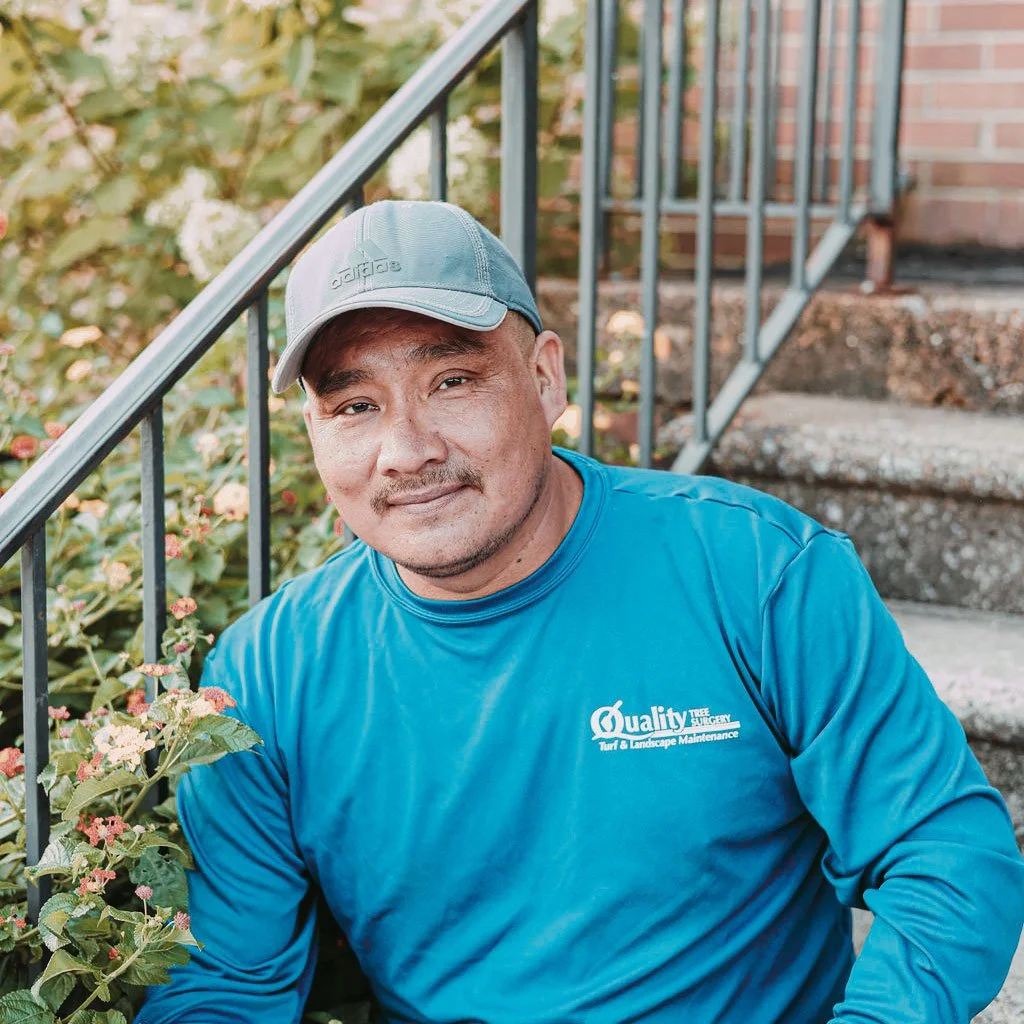 Man in blue long-sleeve shirt and gray cap sitting on stairs outside, near green bushes with pink flowers.