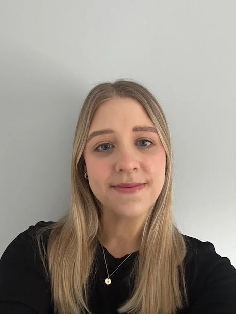 A young woman with long blonde hair, wearing a black top and a necklace, standing against a plain light-colored wall.