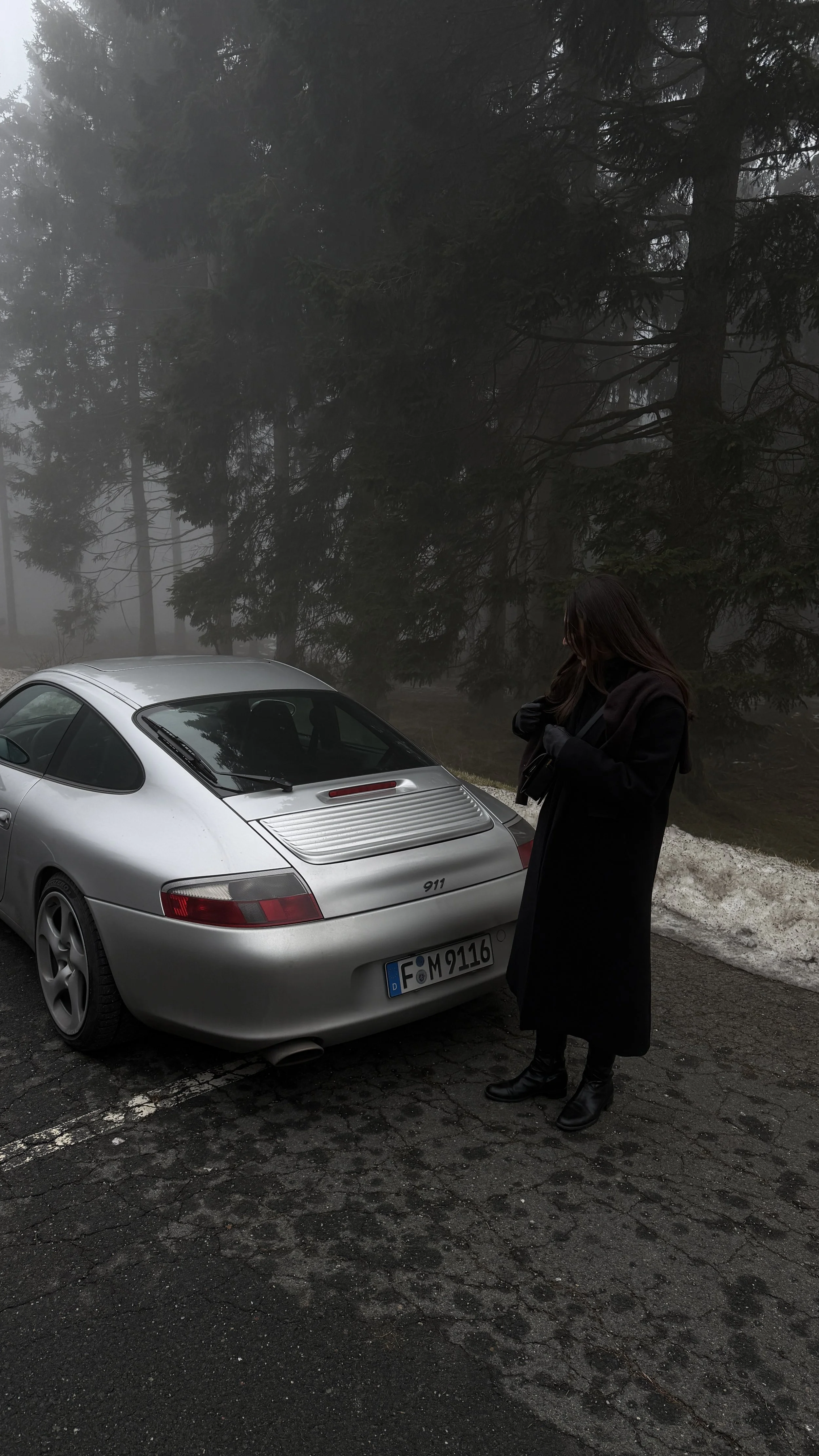 Eine Frau in dunkler Kleidung steht neben einem silbernen Porsche 911 auf einer bewaldeten, nebelverhangenen Straße.