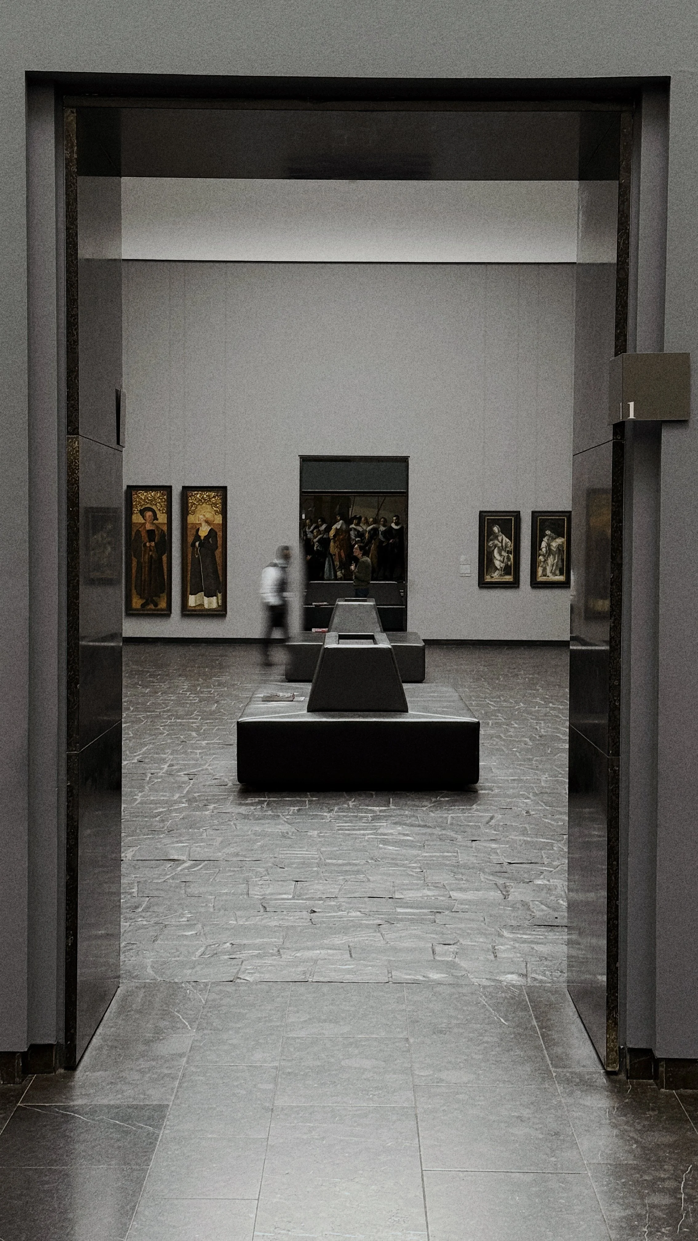 Blick in ein Museum mit Gemälden an den Wänden, Sitzbänken und einer Person, die den Raum betritt.