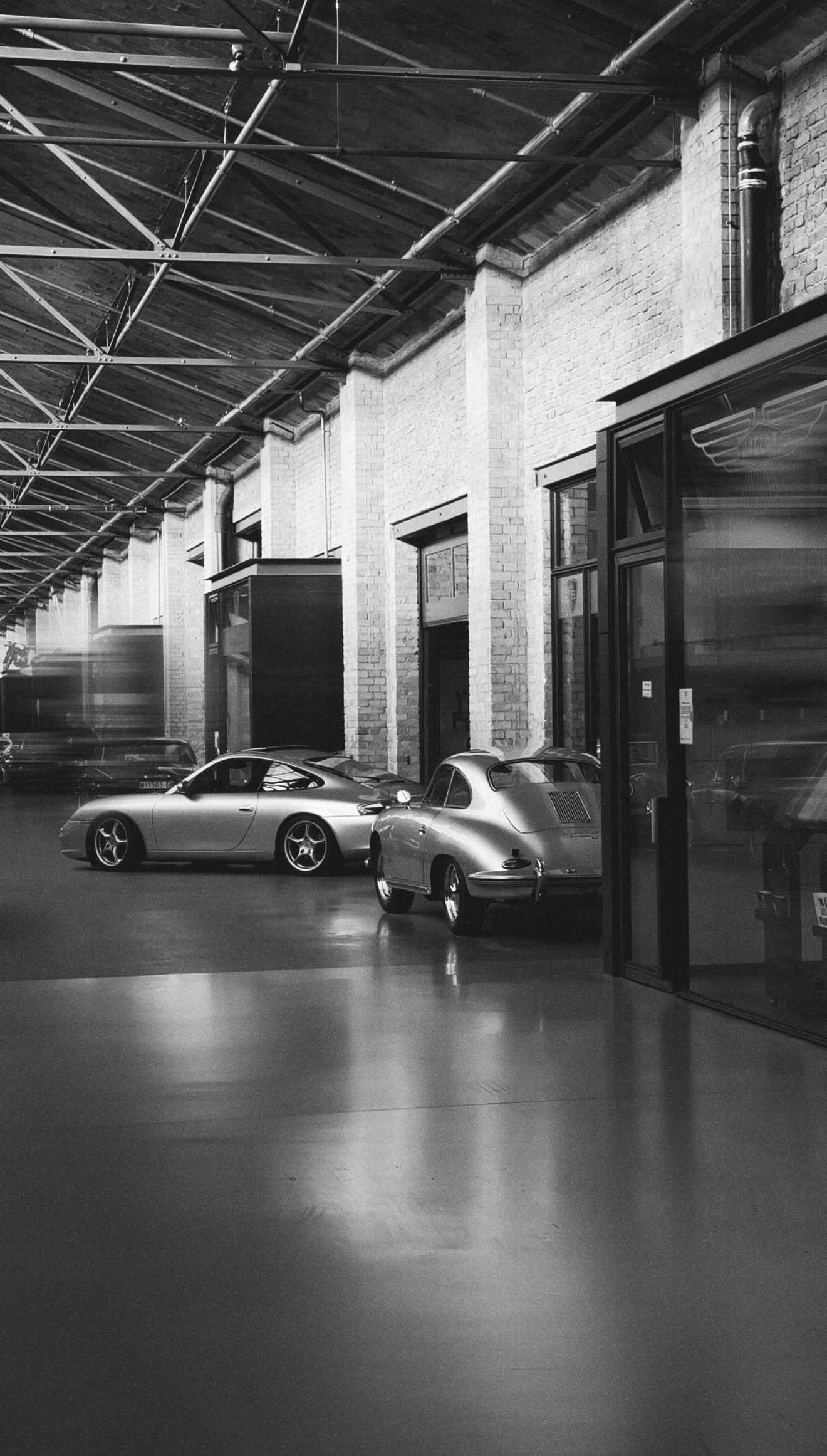 Schwarz-Weiß-Foto eines Garagenhinterhofs mit zwei klassischen Autos, einem modernen Sportwagen, hinter einer Glaswand. Hochsitzdecke und Backsteinwände im Hintergrund.