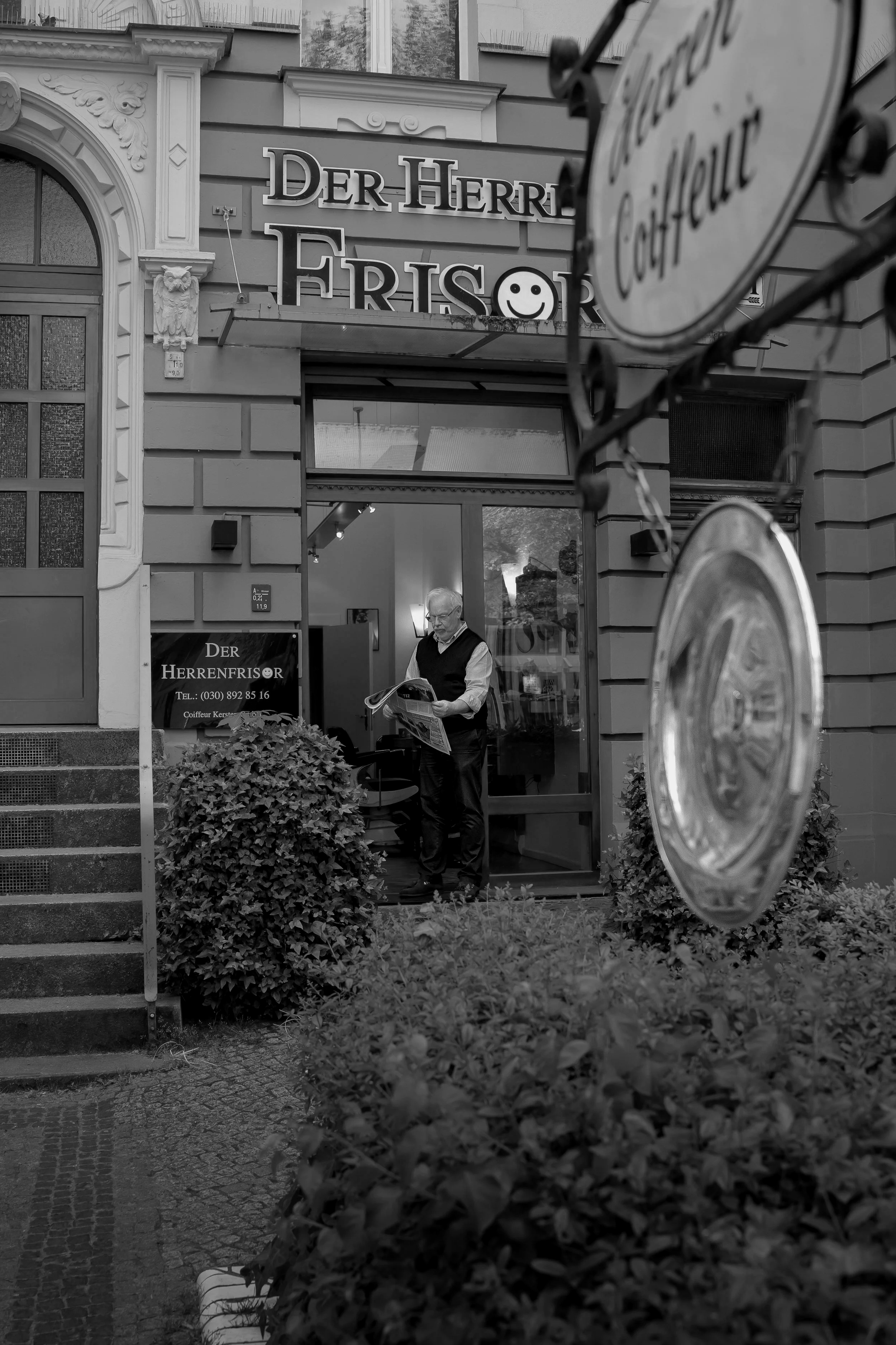 Mann steht vor einem Friseursalon namens "Der Herren Friseur" und liest eine Zeitung, während er die Eingangstür des Ladens betritt oder verlässt. Es ist ein urbanes, mehrstöckiges Gebäude mit klassischen Fassadendetails.