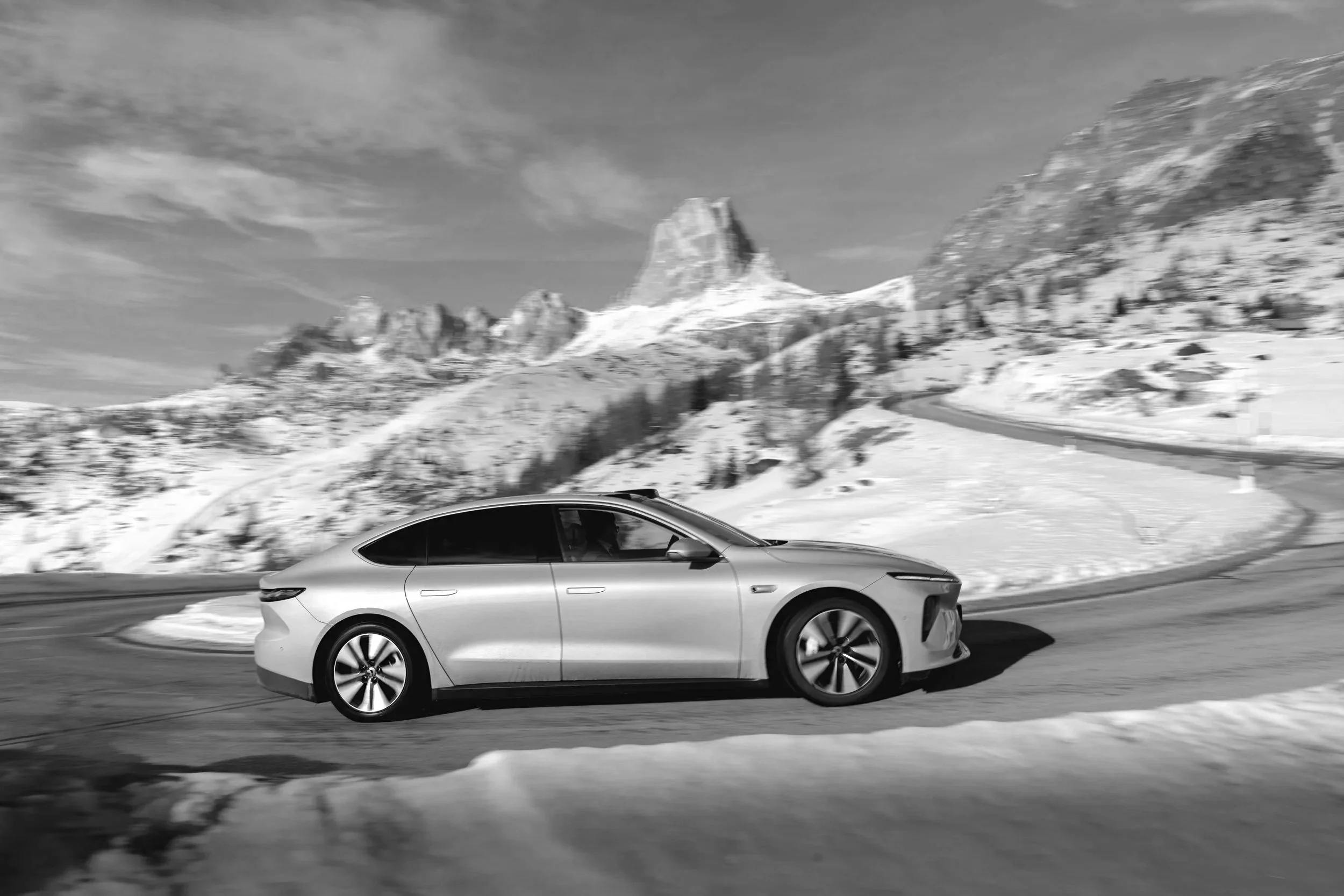 Ein modernes Elektroauto fährt auf einer kurvigen schneebedeckten Straße vor einer winterlichen Bergkulisse in den Bergen, im Hintergrund sind schneebedeckte Gipfel sichtbar, die Szenerie ist winterlich und verschneit.