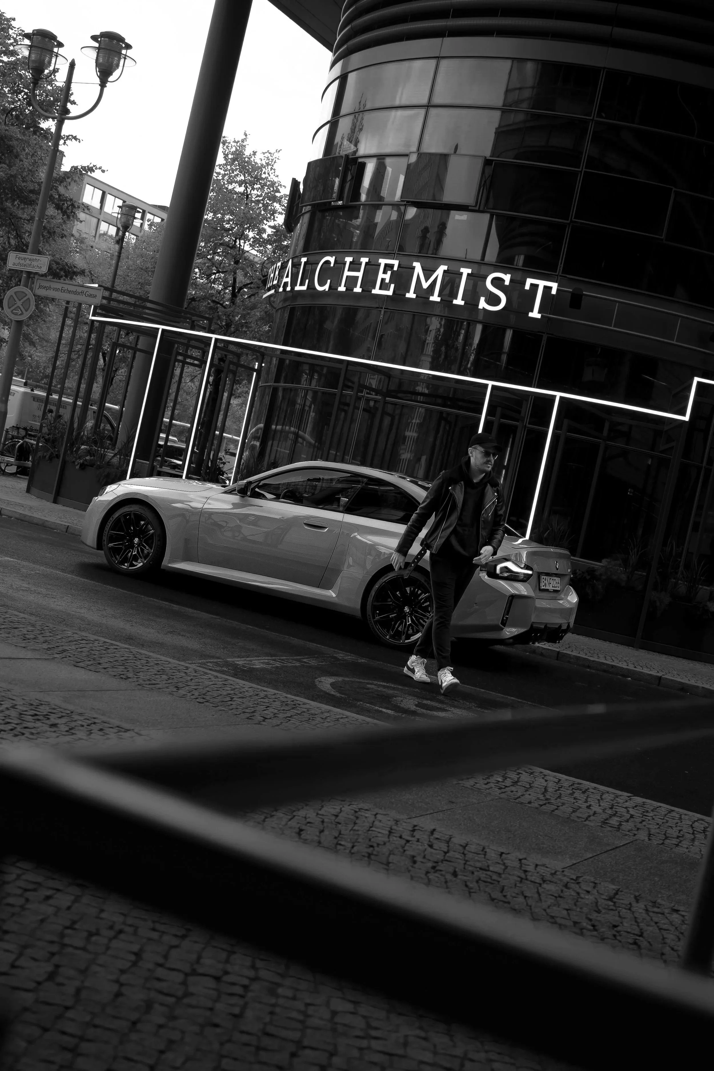 Ein Mann läuft über eine Straße in einer Stadt, im Hintergrund ein modernes schwarzes Gebäude mit roten Neonlichtern und einem großen Schriftzug "ALCHEMIST". Ein graues Auto steht am Straßenrand.
