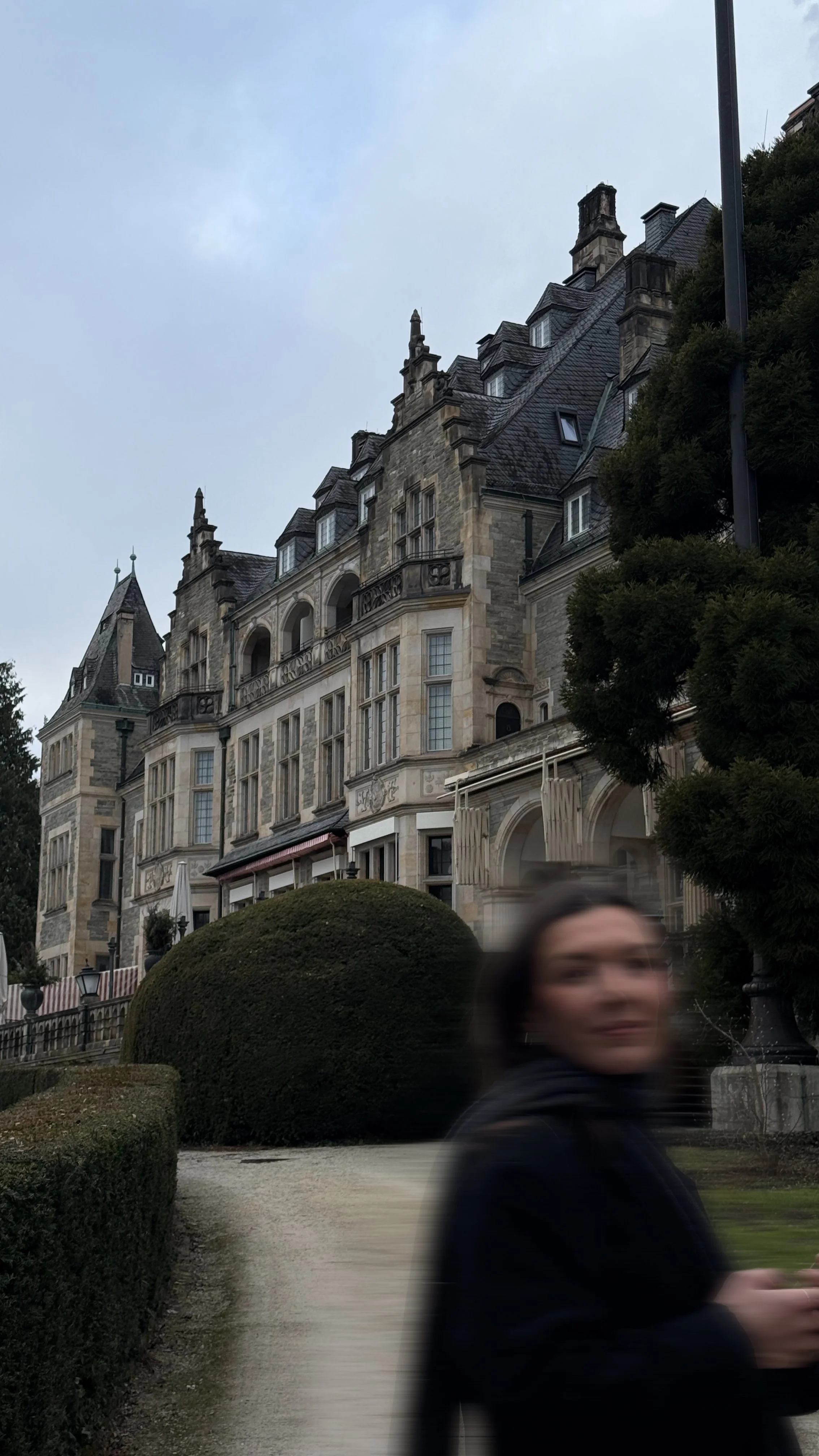 Ein elegantes, historisches Schloss mit gotischer Architektur und mehreren Türmen und Fenstern. Im Vordergrund ist eine verschwommene Person zu sehen, die auf einem Gehweg steht, umgeben von gepflegten Büsche und Bäumen. Der Himmel ist bewölkt.