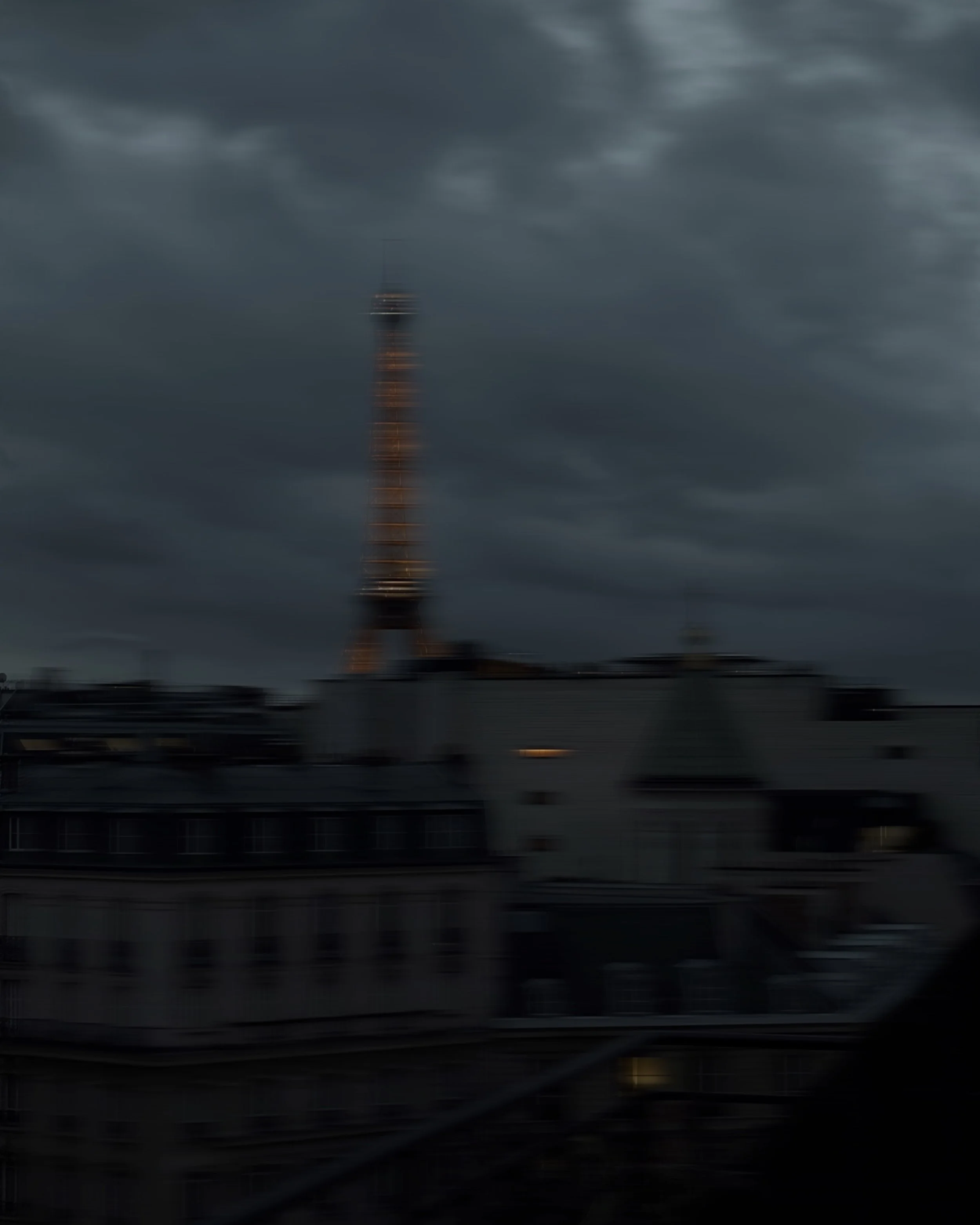 Dach und Türme eines Parisern Gebäudes bei bewölktem Himmel, mit dem Eiffelturm im Hintergrund, unscharf aufgenommen in der Dämmerung.